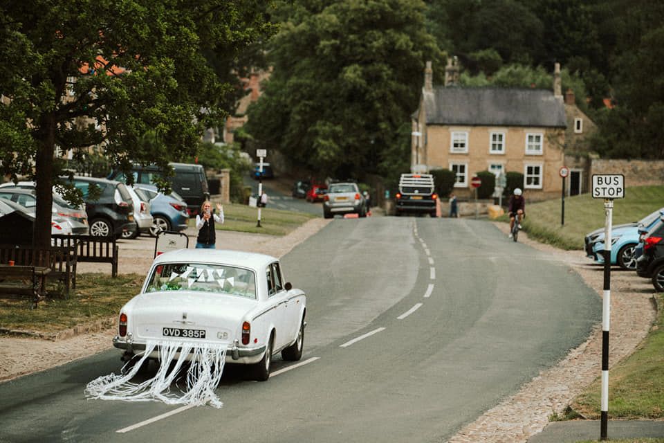 Classic white vintage wedding car with trailing decorations driving down a curved village road.