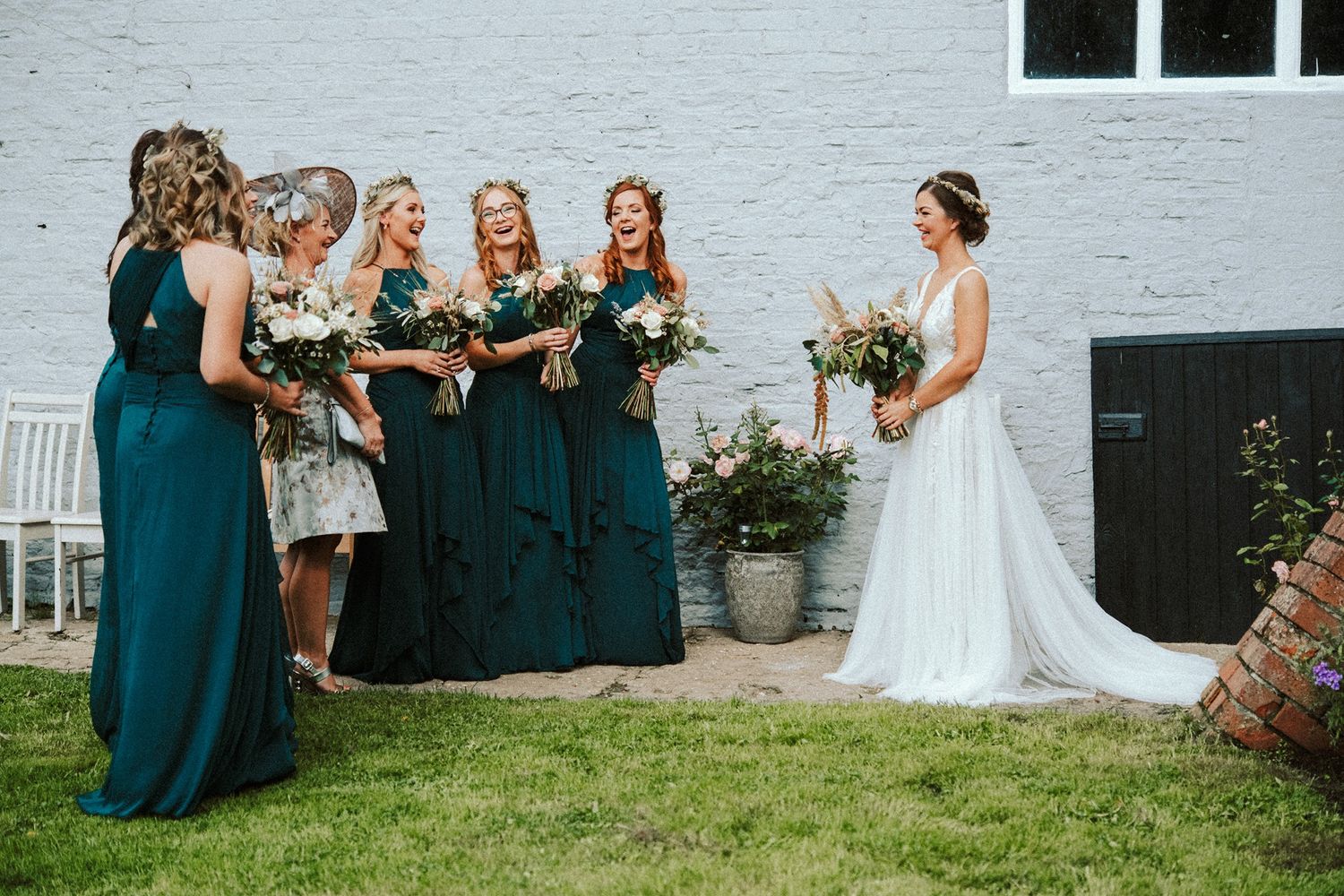 Wedding party in emerald green dresses poses against white brick wall.