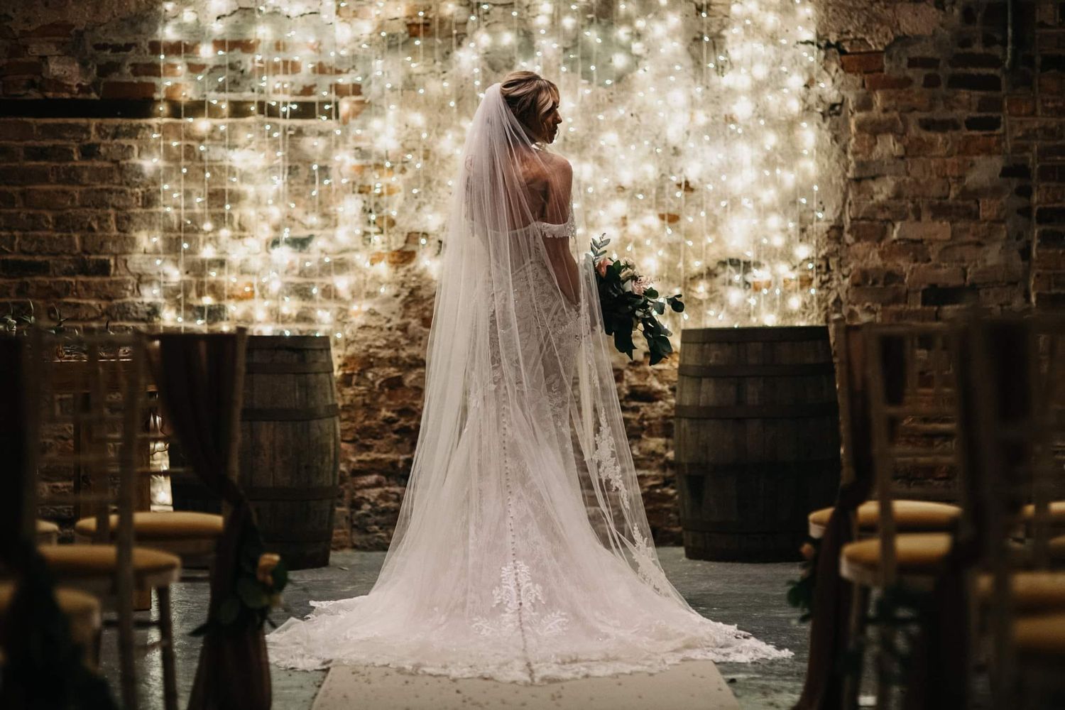 A bride in a long veil poses in front of an illuminated brick wall with barrels.