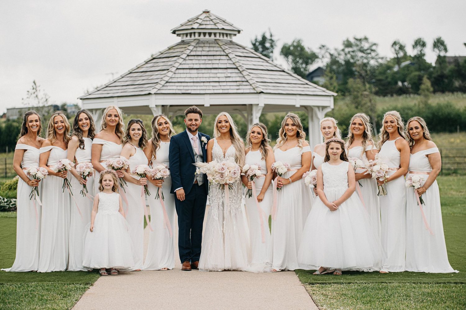 Wedding party poses in white dresses against a white gazebo backdrop.