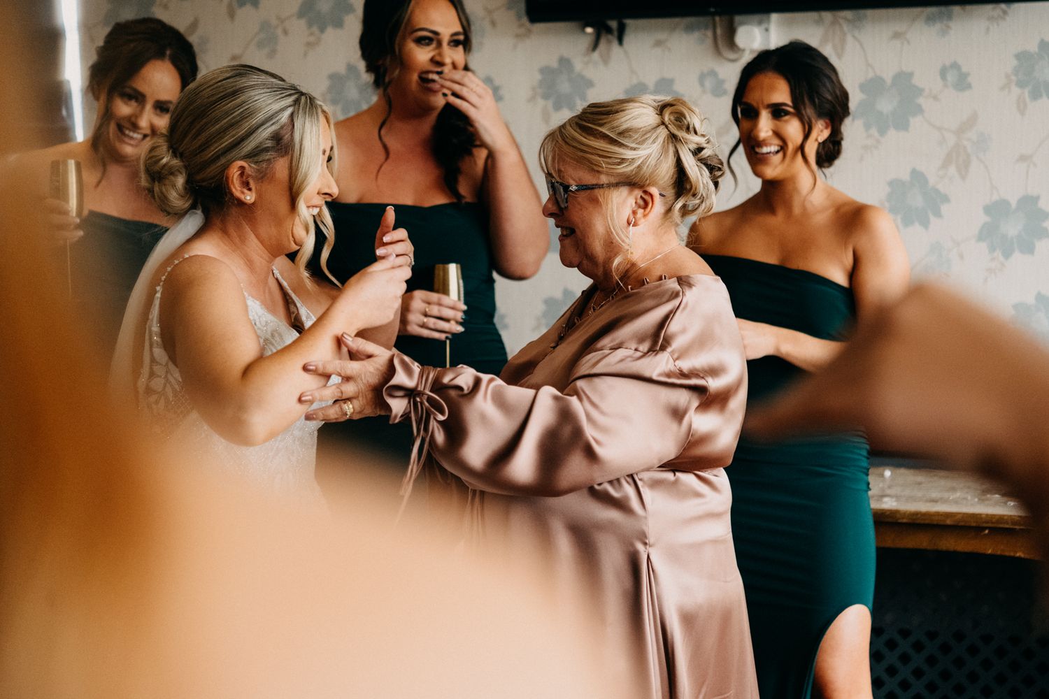 A group getting ready in a vintage-wallpapered room with one wearing a blush robe and others in dark dresses.