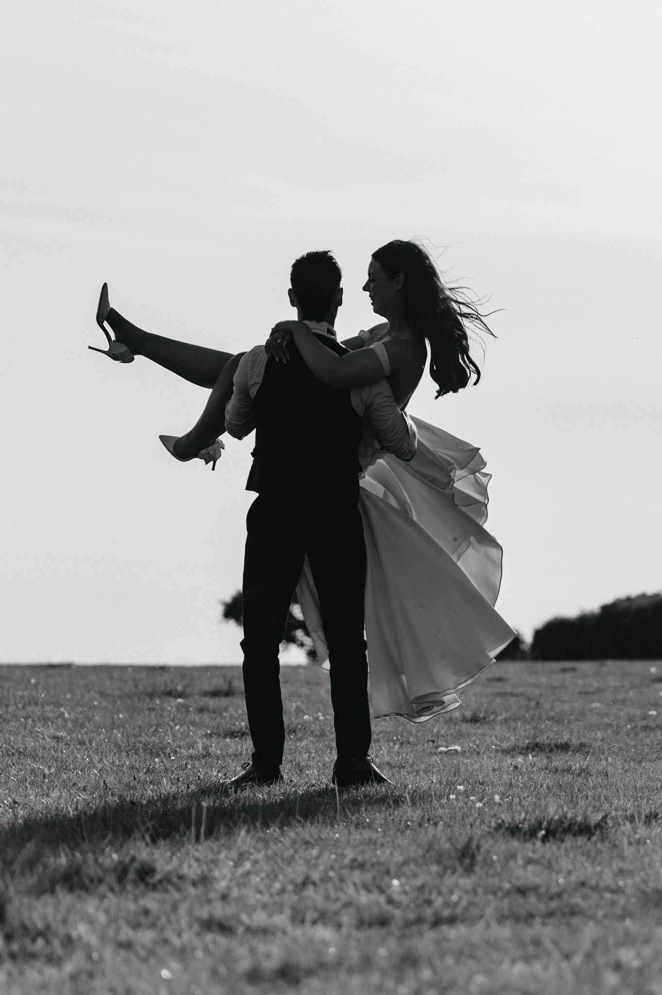 Black and white photo of a groom lifting his bride in a field, her dress flowing in the wind.
