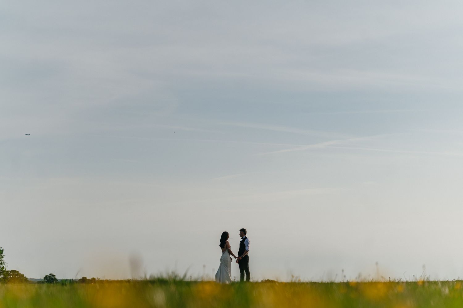 Couple standing together in a vast open field at dusk, silhouetted against a soft blue sky.
