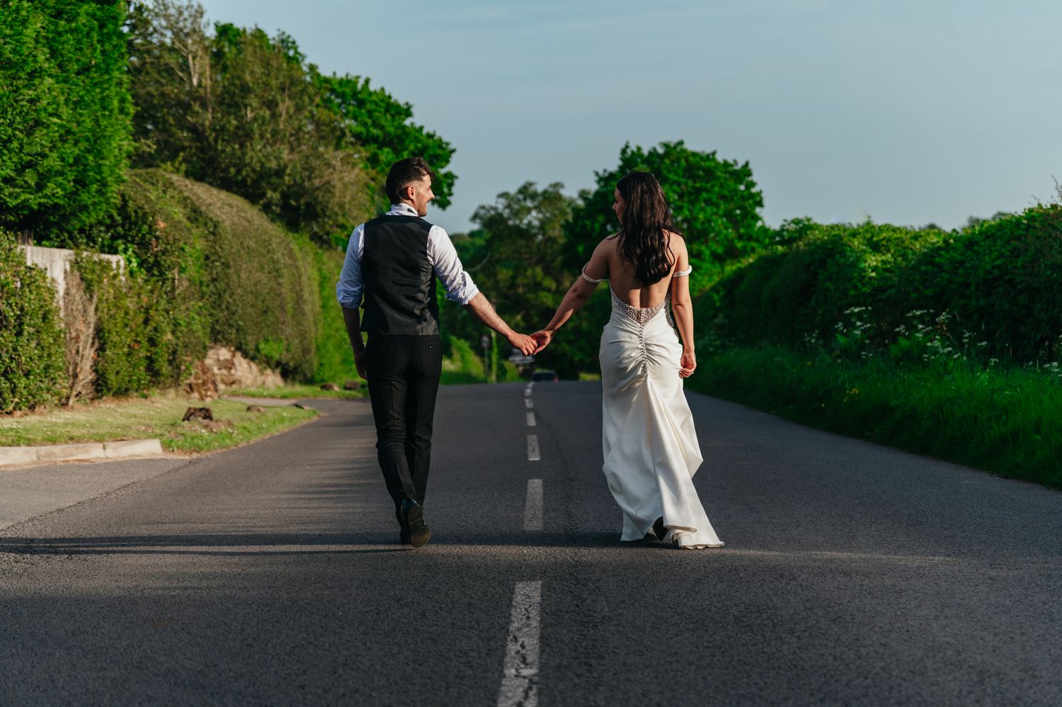 Bride and groom walking hand in hand down a scenic road lined with green trees on their wedding day.