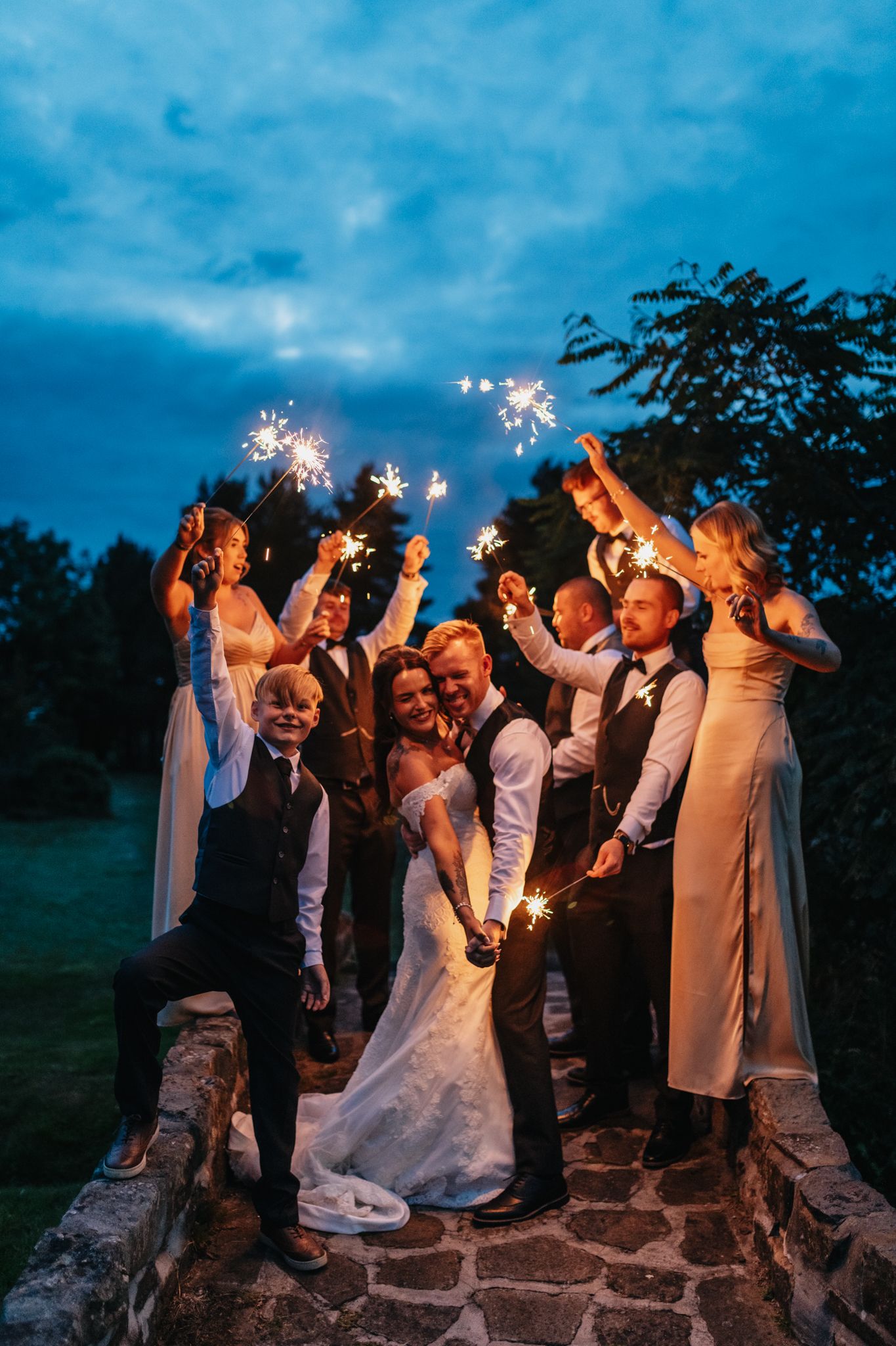 Wedding party celebrates with sparklers at dusk on stone wall beneath moody blue sky.