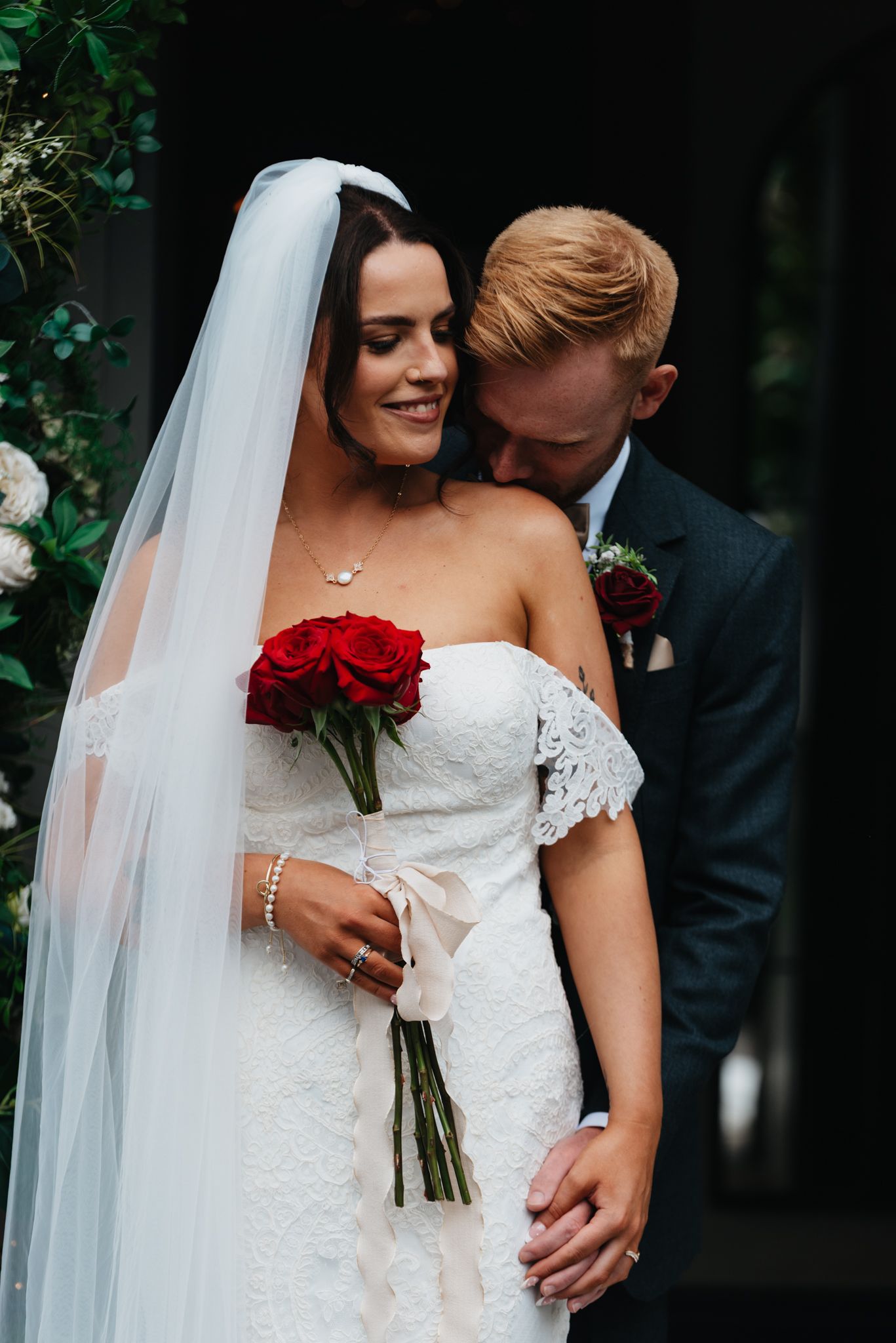 Couple poses intimately on their wedding day with red roses and a flowing white veil against dark foliage.