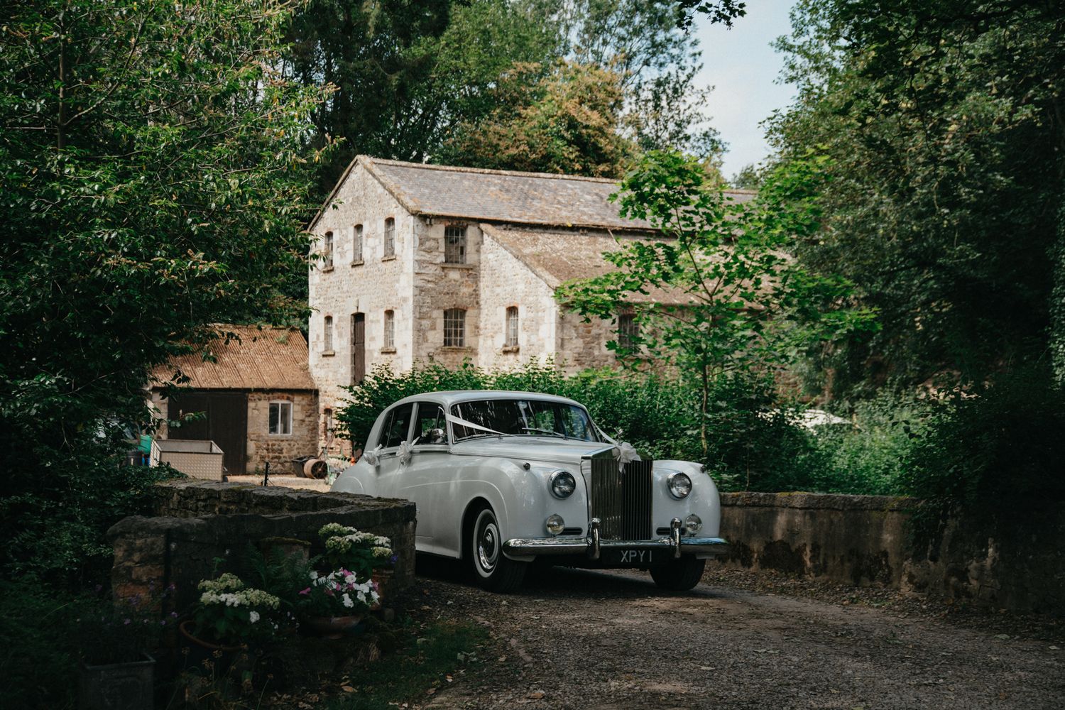 Vintage white Rolls Royce wedding car parked outside rustic stone country house surrounded by green trees.