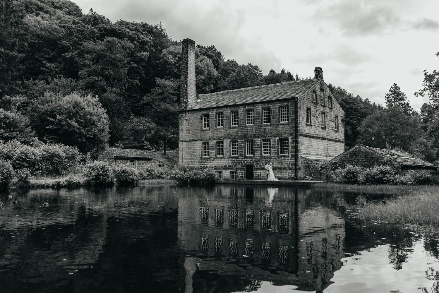 Historic brick mill building reflects in still water surrounded by lush trees in black and white landscape photography.