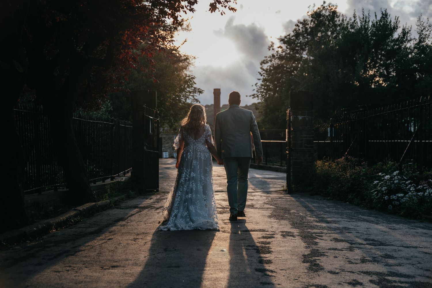 Two newlyweds walk hand in hand on sunlit path during sunset with long shadows cast on ground behind them.
