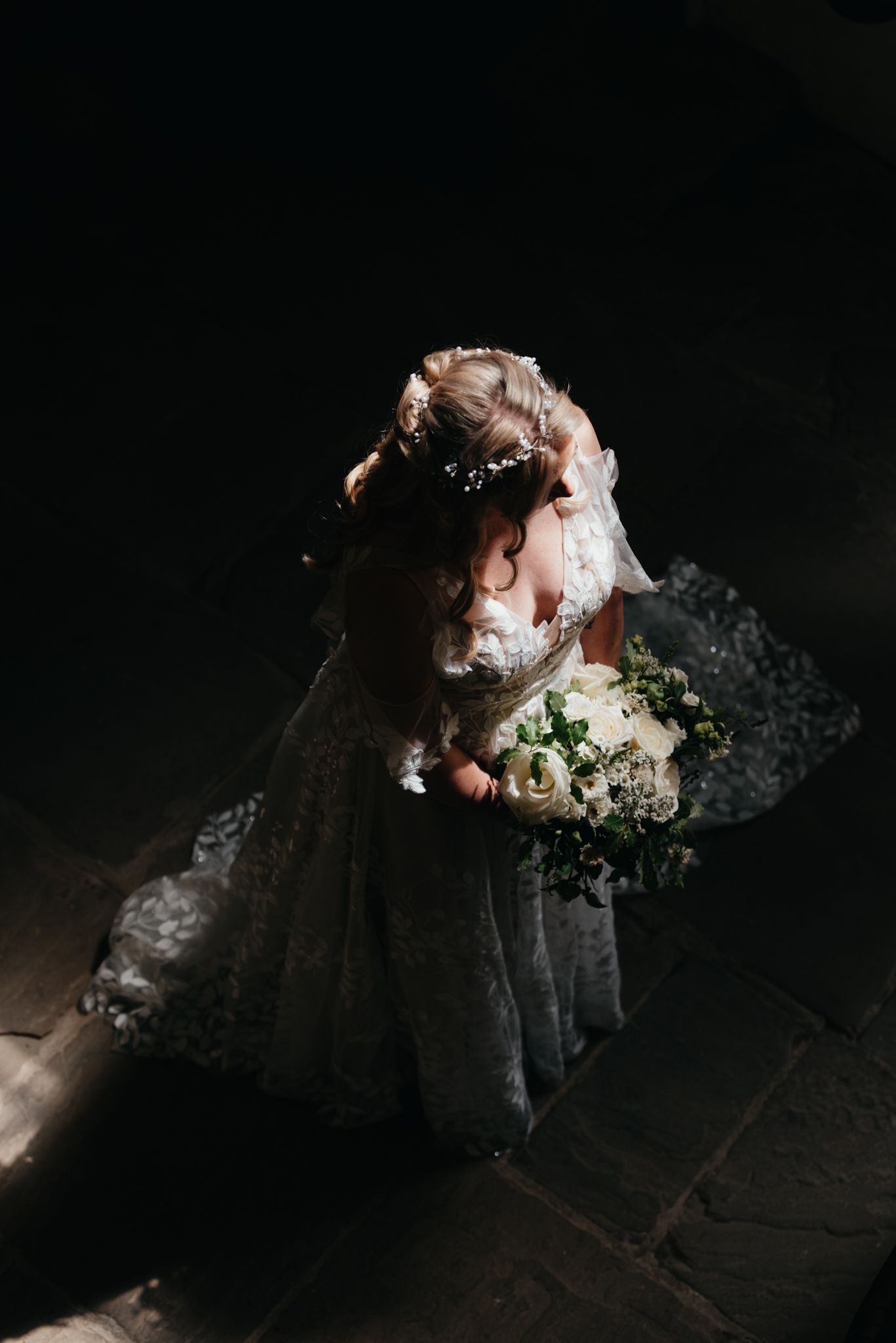 A bride in white lace gown and veil holds a romantic bouquet against a dark background in moody lighting.