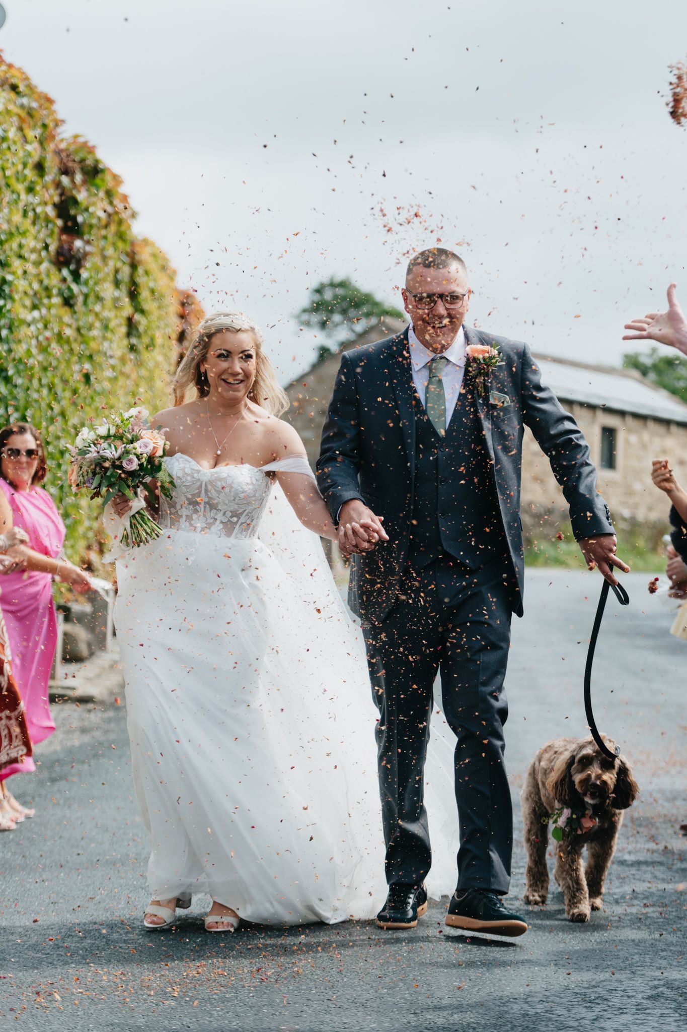 A joyful wedding couple walks down a road with their dog while confetti falls around them on their special day.