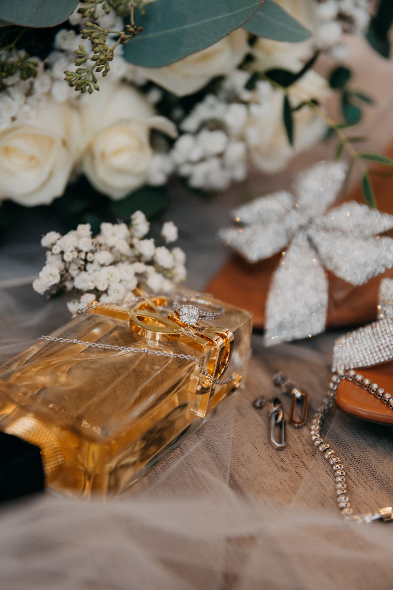 Wedding accessories including perfume bottle, white roses, jewelry and a decorative bow arranged on a textured surface.