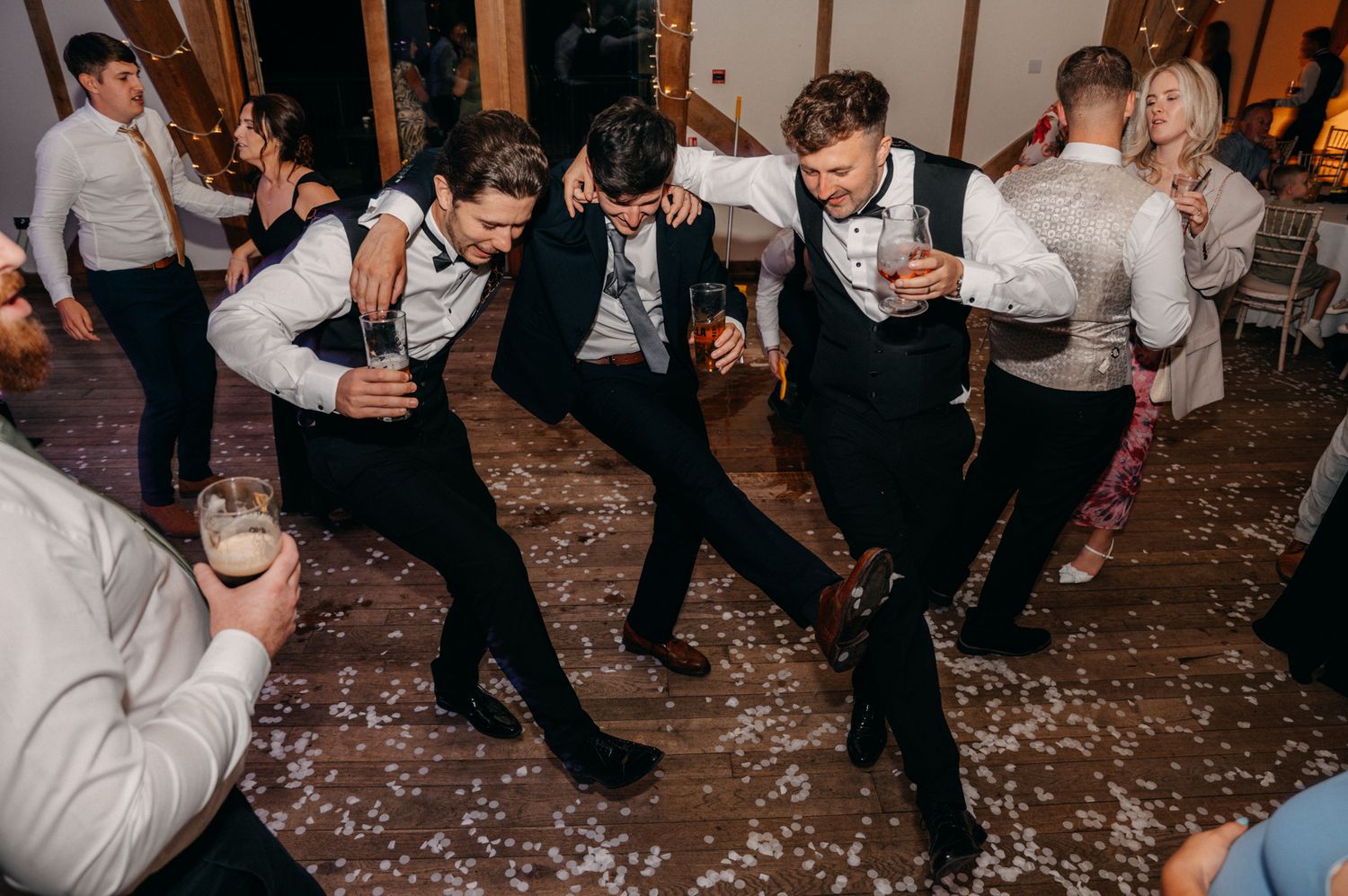 A crowd of wedding guests dancing and celebrating on a confetti-covered dance floor at a reception party.