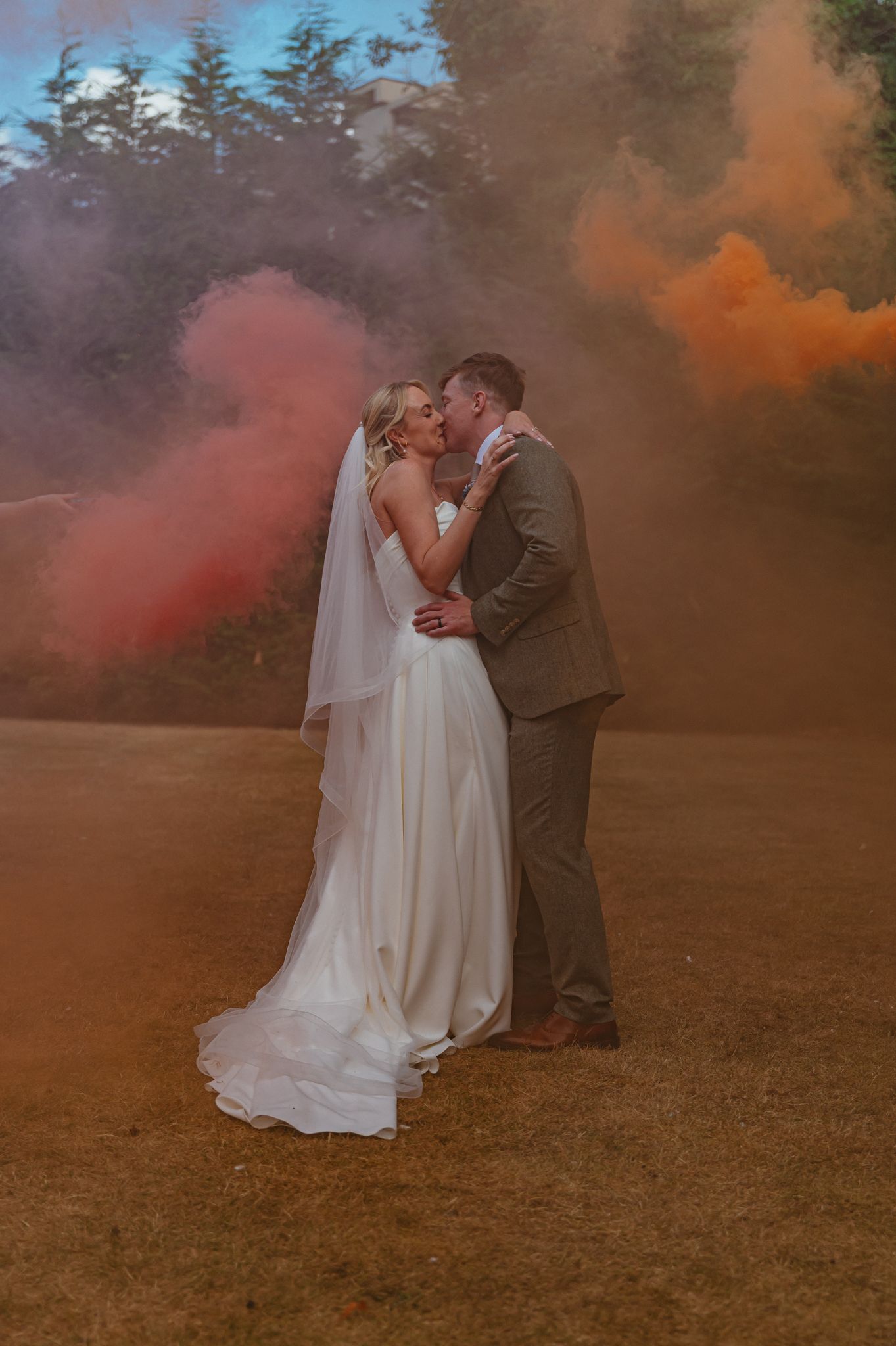 Bride and groom kissing surrounded by colorful orange and pink smoke bombs outdoors at their wedding celebration.