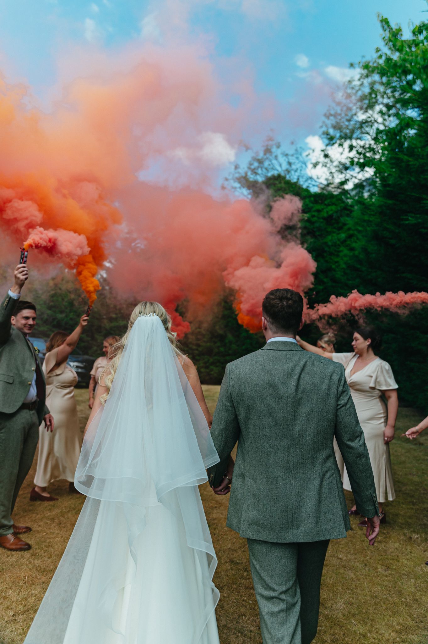 A bride and groom walk through colorful orange and pink smoke bombs held by wedding guests outdoors.