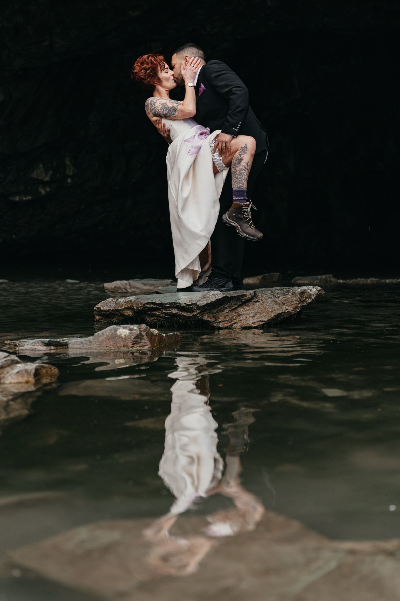 Groom lifts bride in wedding attire on rocks by water, sharing a romantic kiss with their reflection visible below.