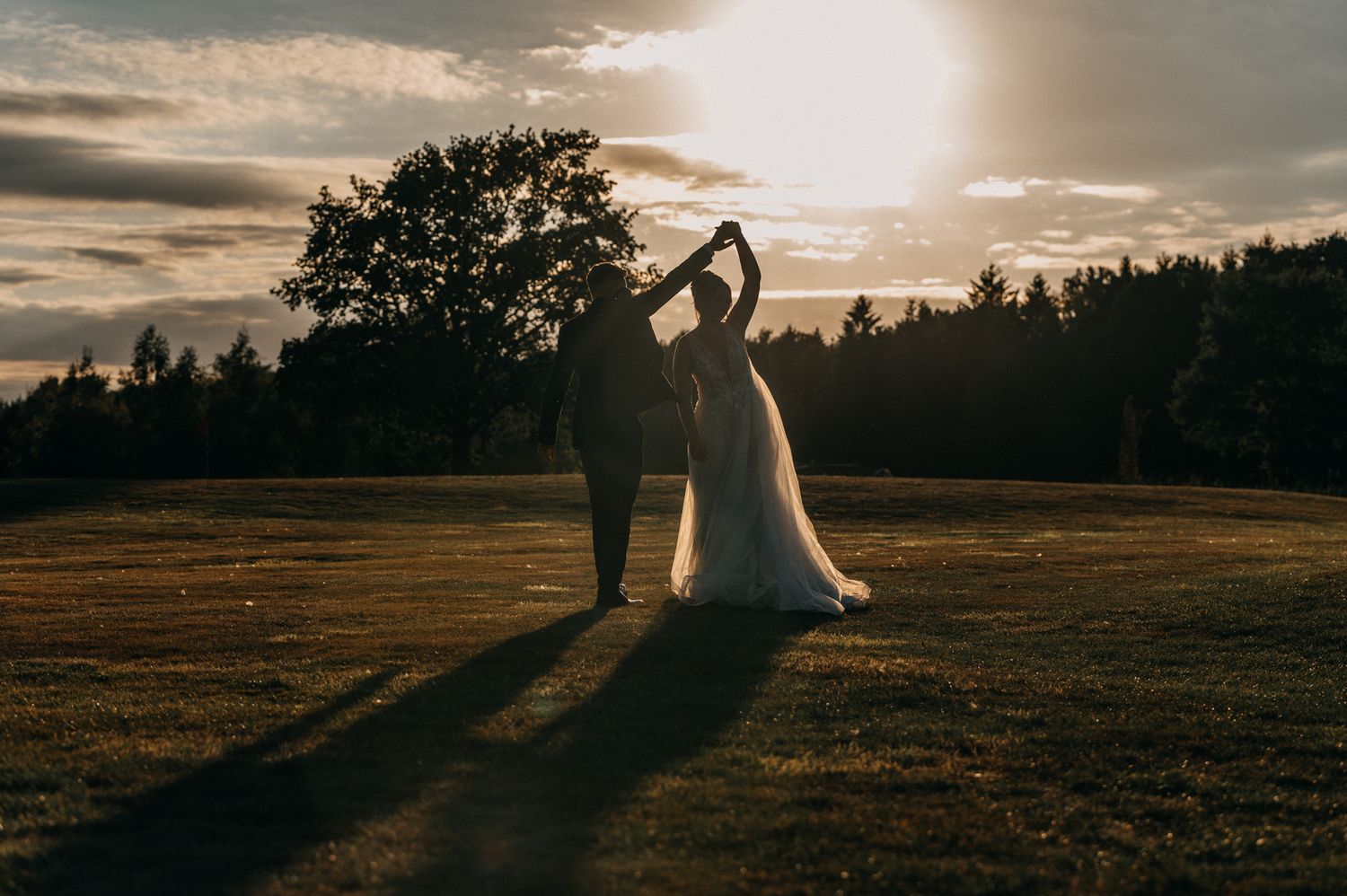 A romantic sunset silhouette of newlyweds standing in a golden field as the sun casts long shadows through the trees.