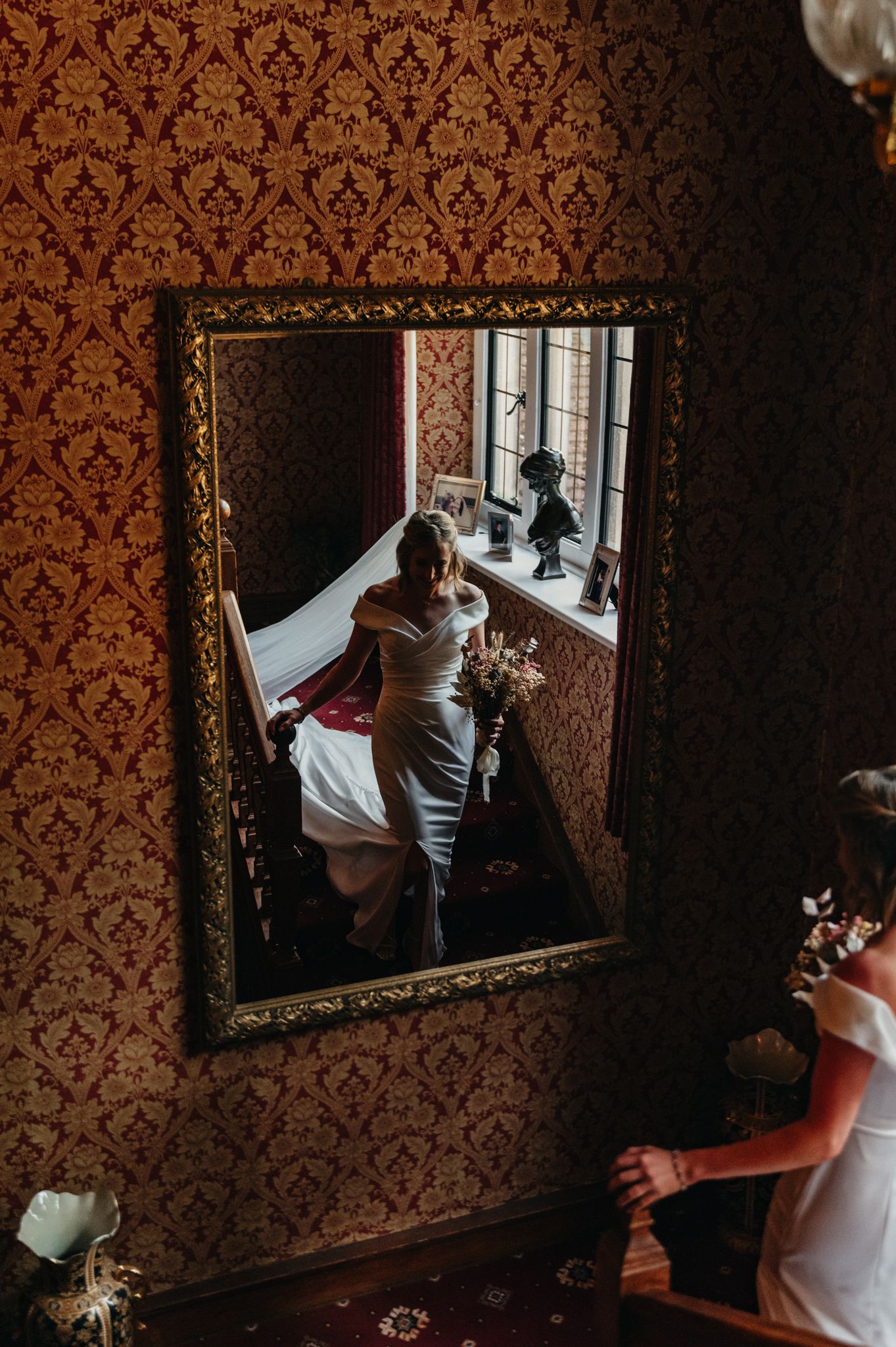 Bride in white gown reflected in ornate gold mirror on red damask wall, ascending staircase with bouquet.
