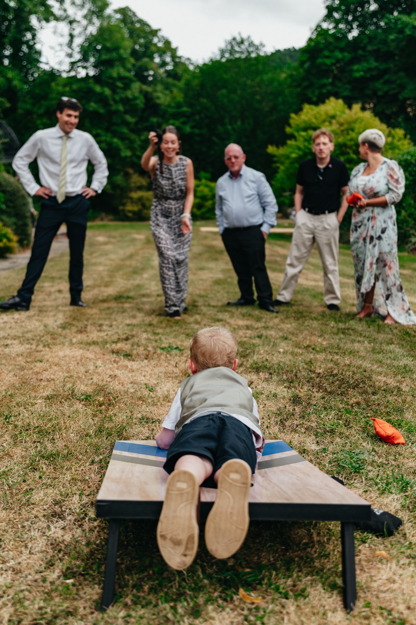 Young child lying on a bench at an outdoor wedding reception while guests socialize on a green lawn.