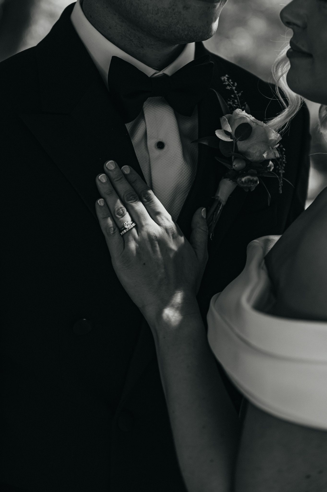 Black and white wedding photo of bride and groom embracing, showcasing diamond ring and boutonniere.