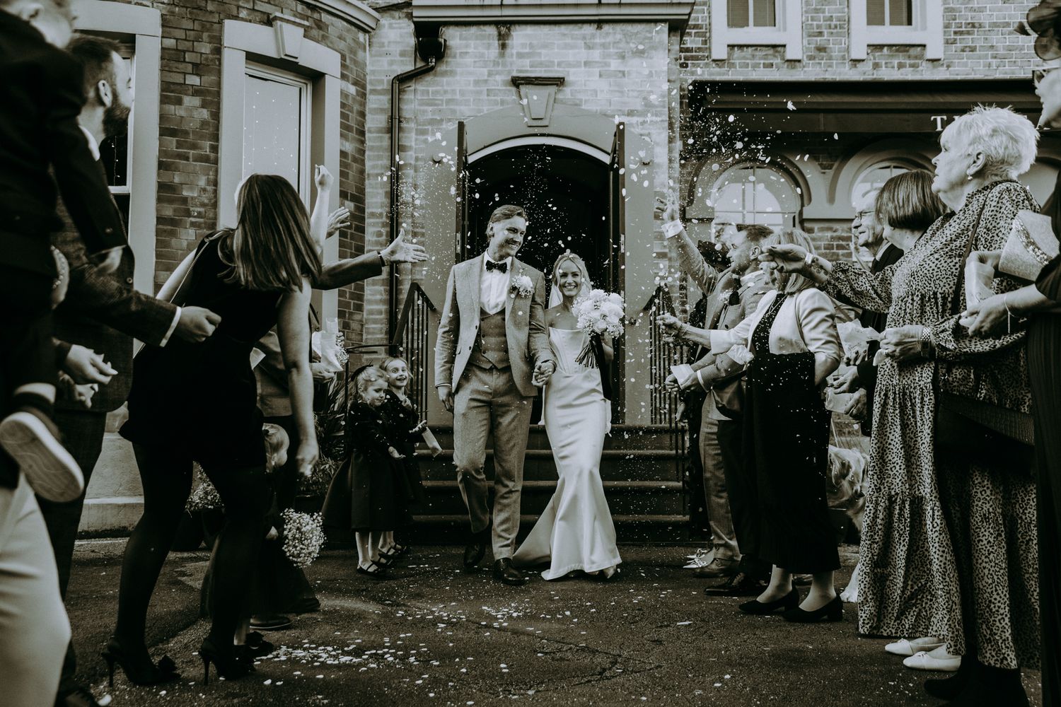 Wedding guests throw confetti on newlyweds exiting ornate stone building during joyful outdoor celebration. Faversham, Leeds