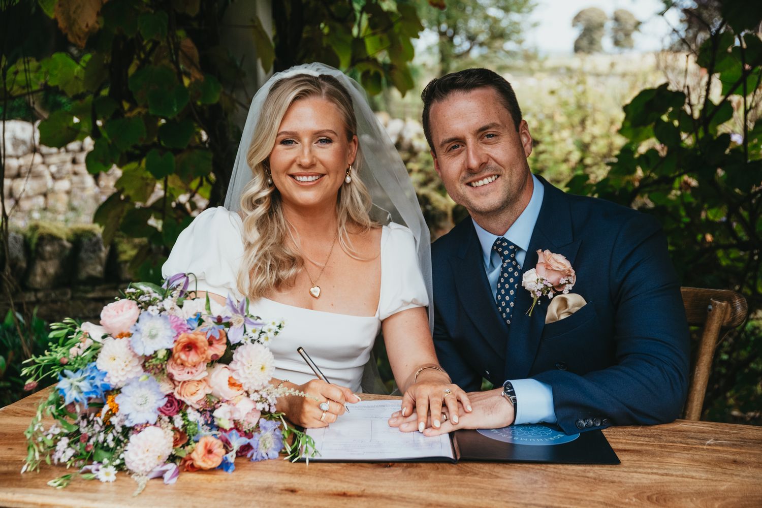 Wedding couple sits at rustic wooden table with colorful floral arrangements.