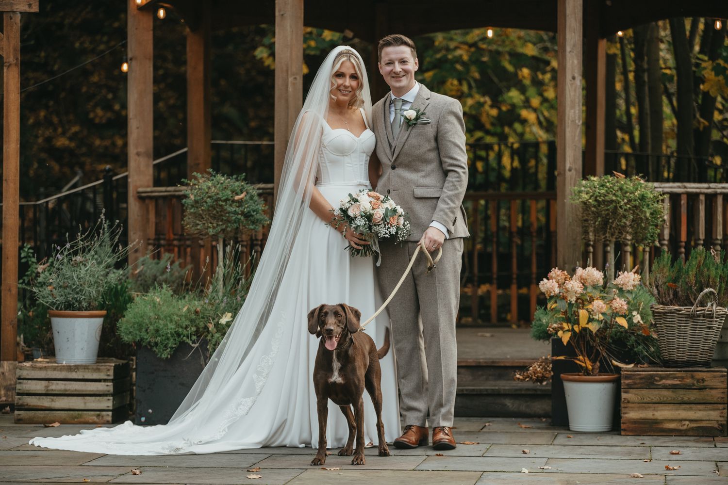 A newlywed couple stands with their brown dog on a rustic wooden deck decorated with potted plants and flowers. The Venue, Halifax