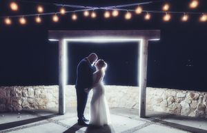 Couple sharing romantic kiss under glowing arch with string lights at night wedding ceremony.