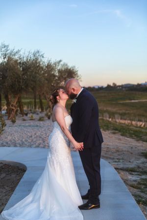 Bride and groom share a romantic kiss at sunset on a garden path, bride in white gown, groom in dark suit.