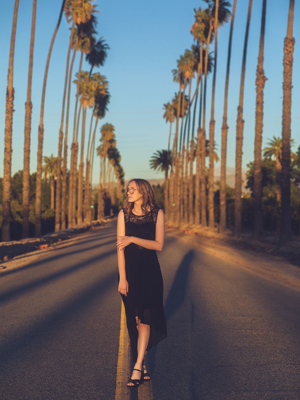 Woman in black dress standing on palm tree-lined road at golden hour sunset.