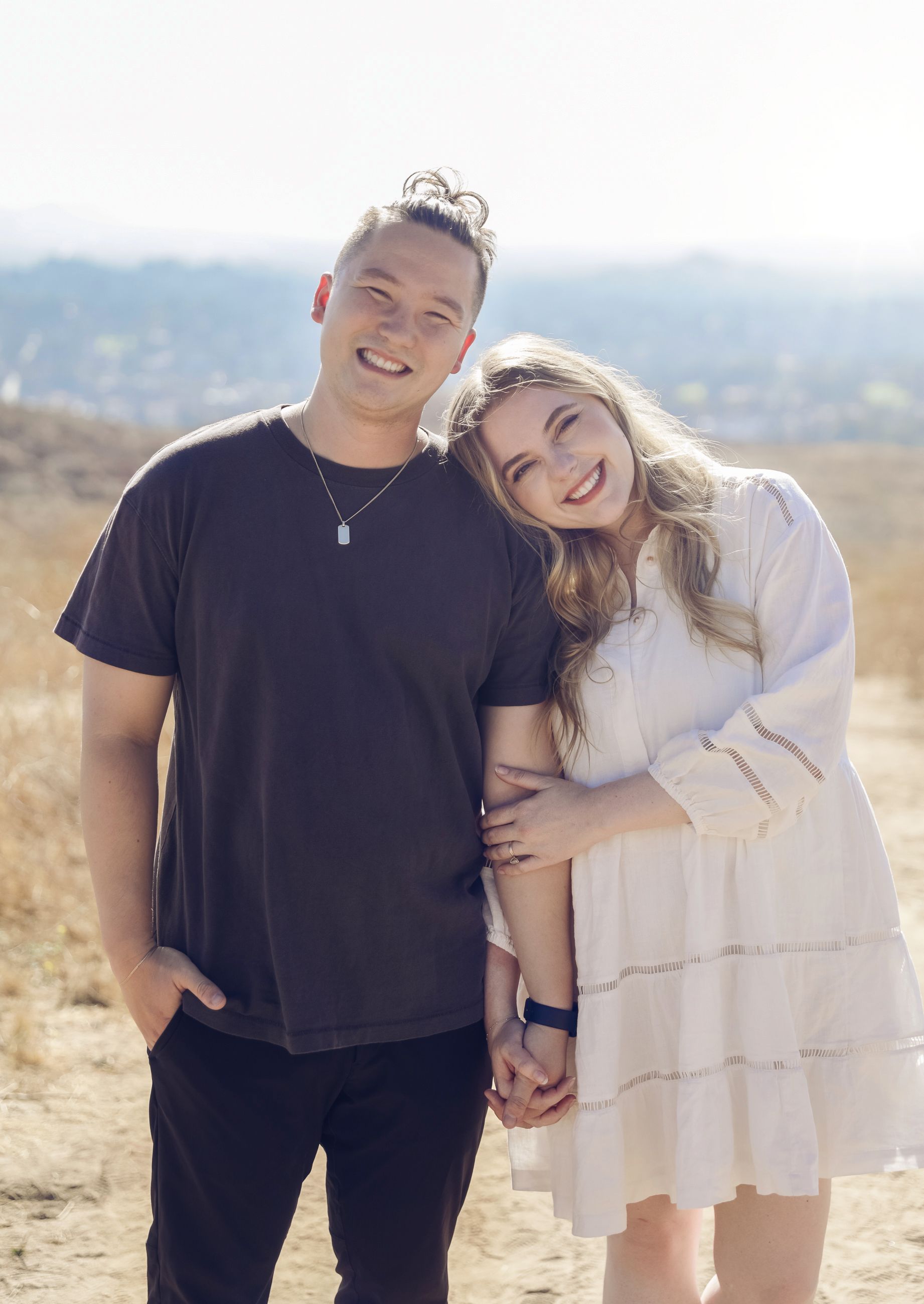 A happy couple poses together outdoors on a sunny day, with one wearing black and the other in a light-colored dress.