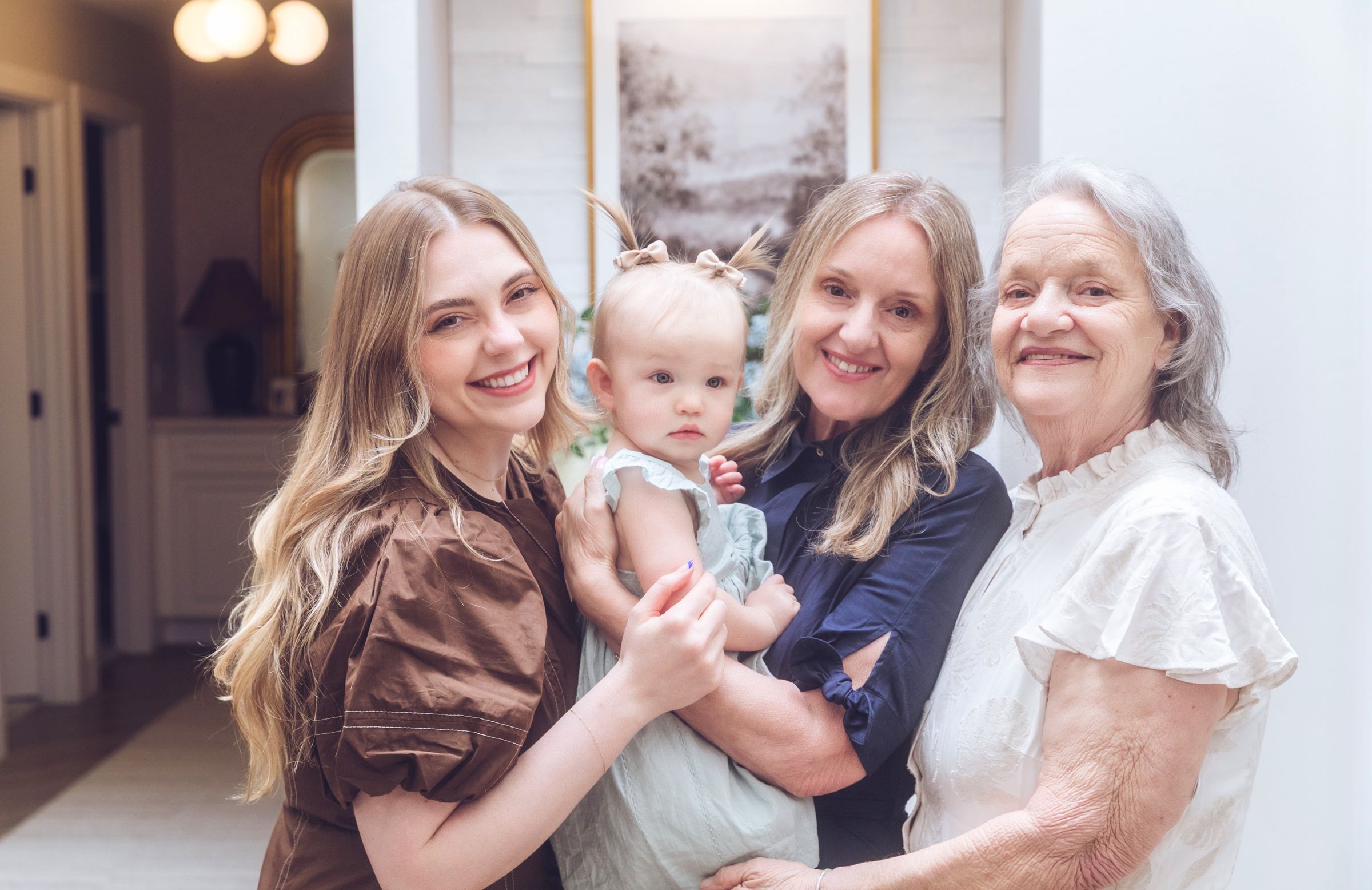 Four generations of women smile together, with a young woman holding a baby flanked by two older relatives indoors.
