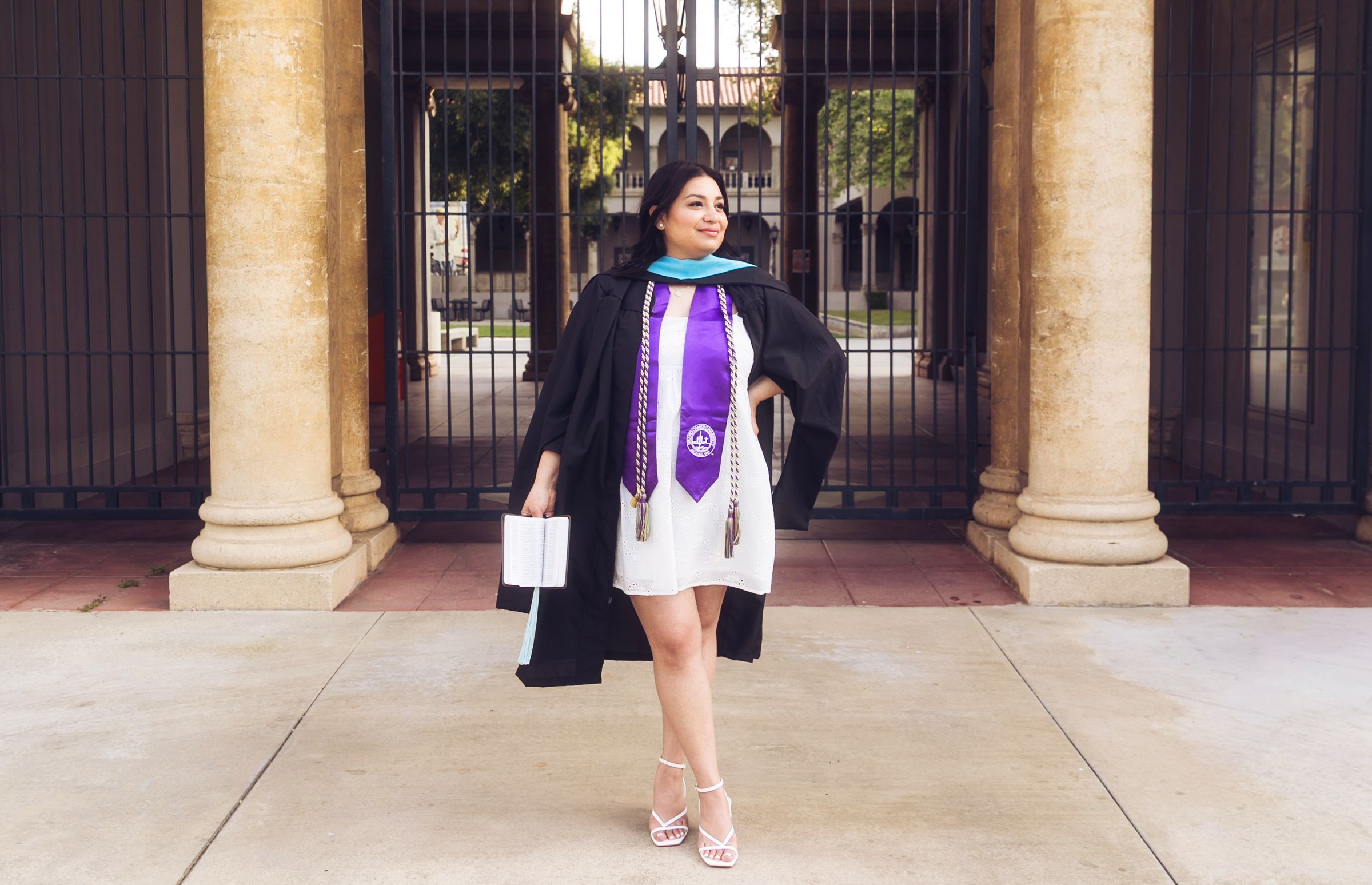 A woman in black graduation gown with purple sash holds cap while posing at university entrance columns.