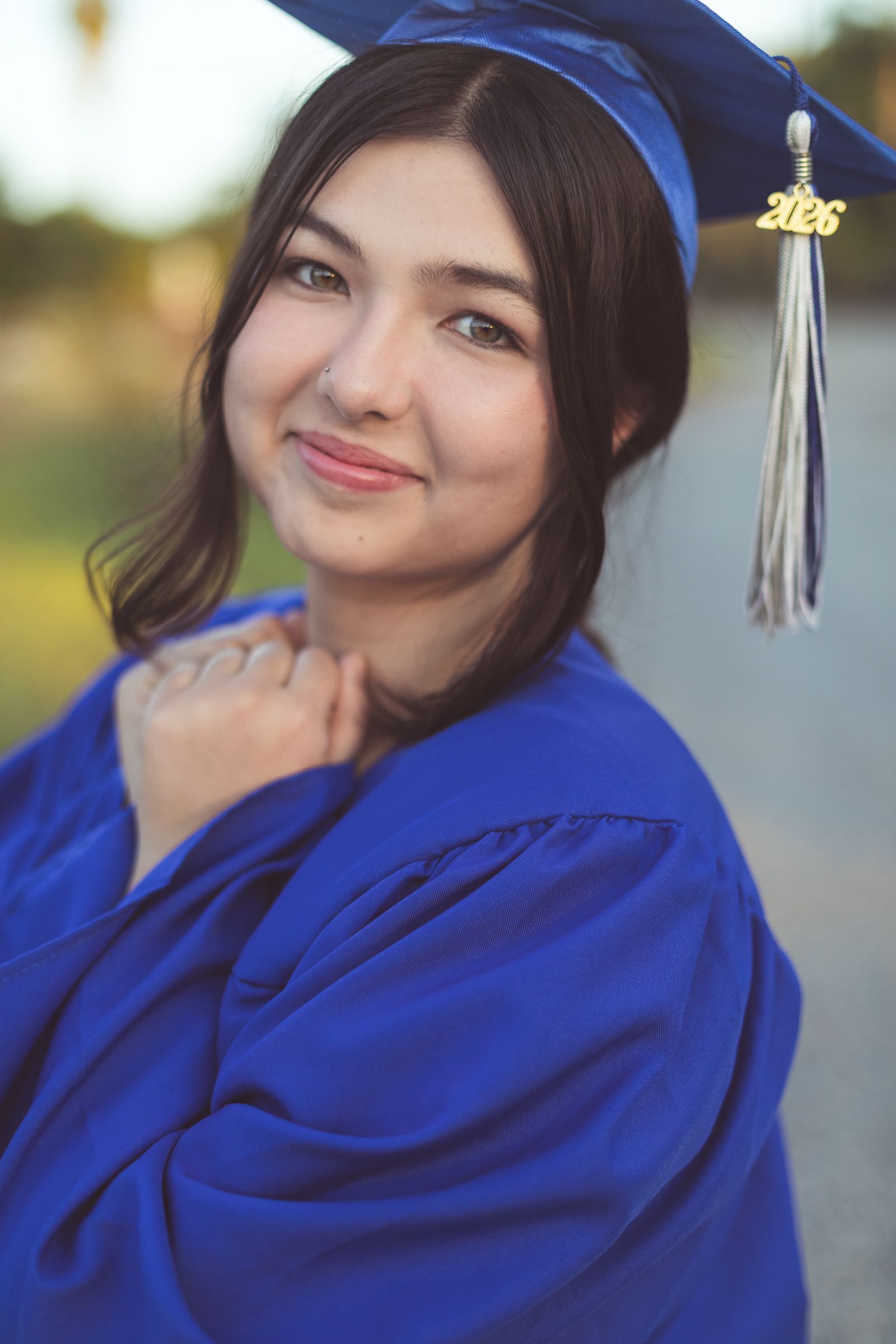 Young woman in blue graduation cap and gown with 2026 tassel smiles confidently outdoors in a portrait photo.