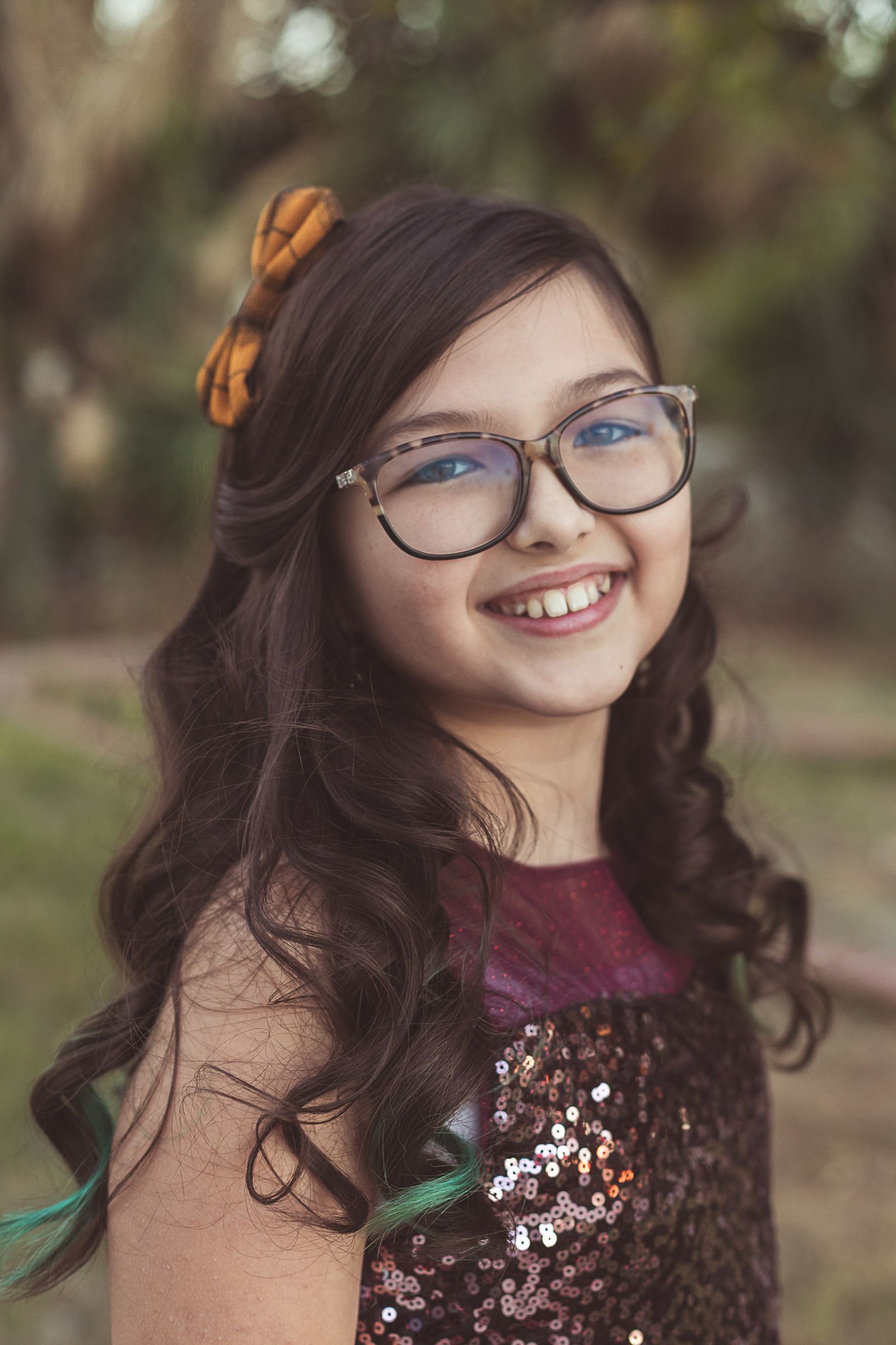 Smiling young girl with long dark hair, glasses, and a flower accessory wearing a sequined dress outdoors.