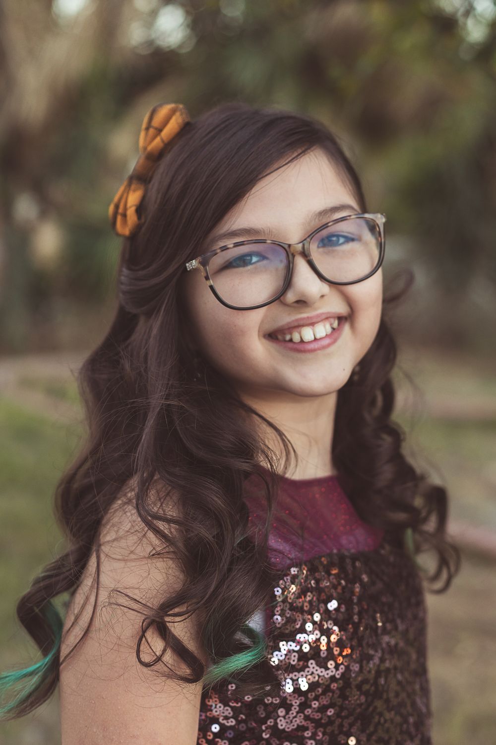 Smiling young girl with long dark hair, glasses, and a flower accessory wearing a sequined dress outdoors.