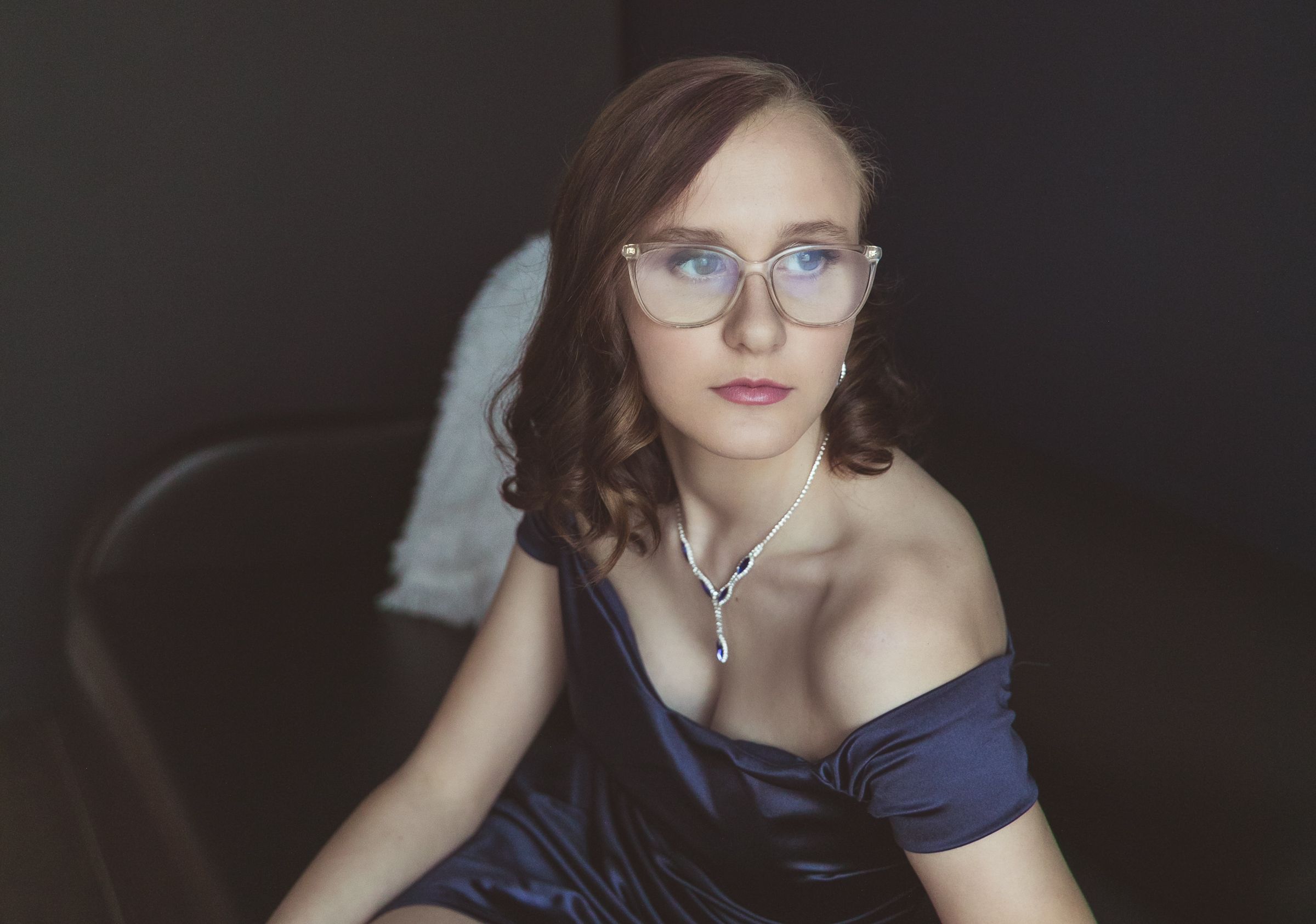 Young woman in blue off-shoulder dress and glasses wearing a silver necklace poses elegantly in a dimly lit room.