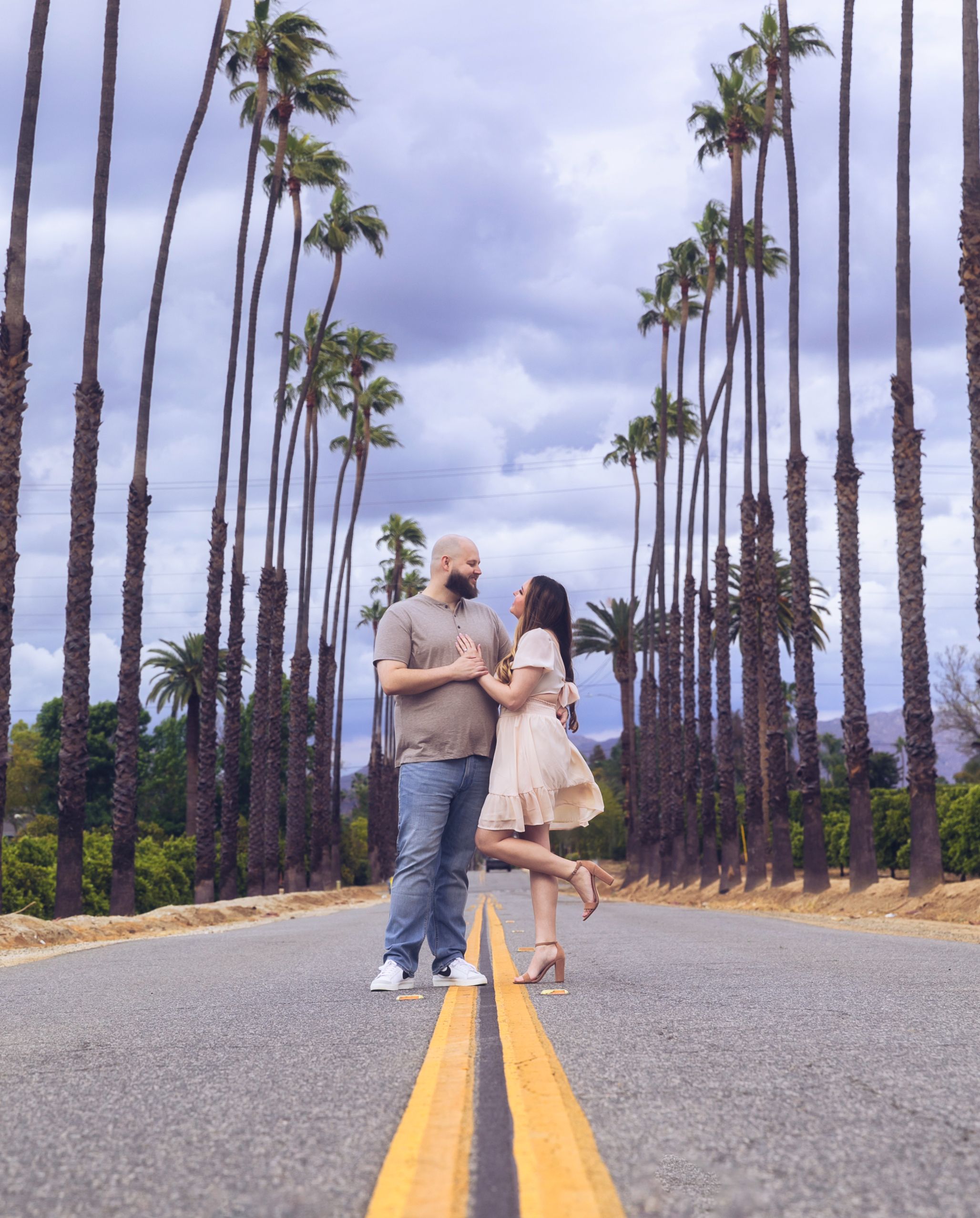 Couple standing on a palm tree-lined road under dramatic cloudy skies, sharing a romantic moment in Los Angeles.