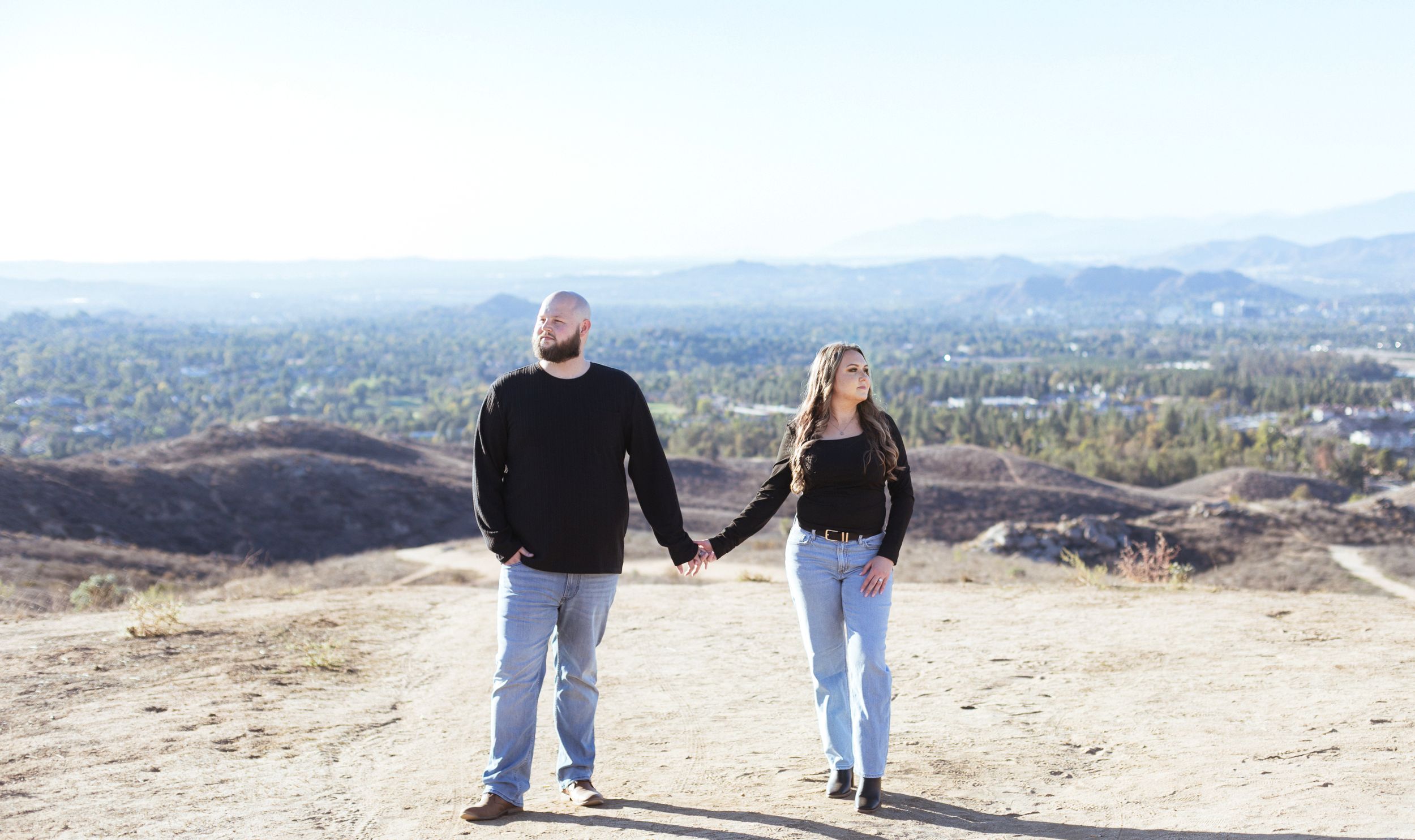 Two people walking along a mountain trail with panoramic city views in the background on a sunny day.