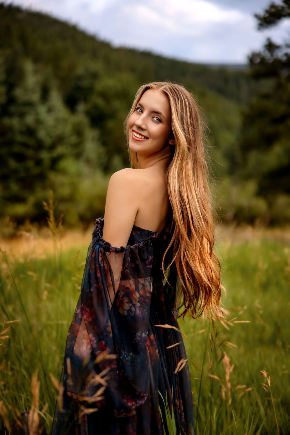 Long-haired woman in floral off-shoulder dress poses in a mountain meadow with evergreen trees in background.
