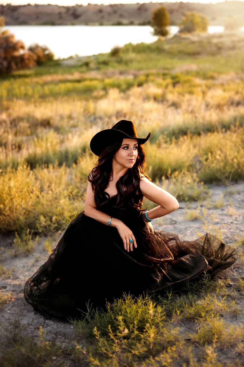Woman in black dress and hat lounges gracefully in golden field at sunset.