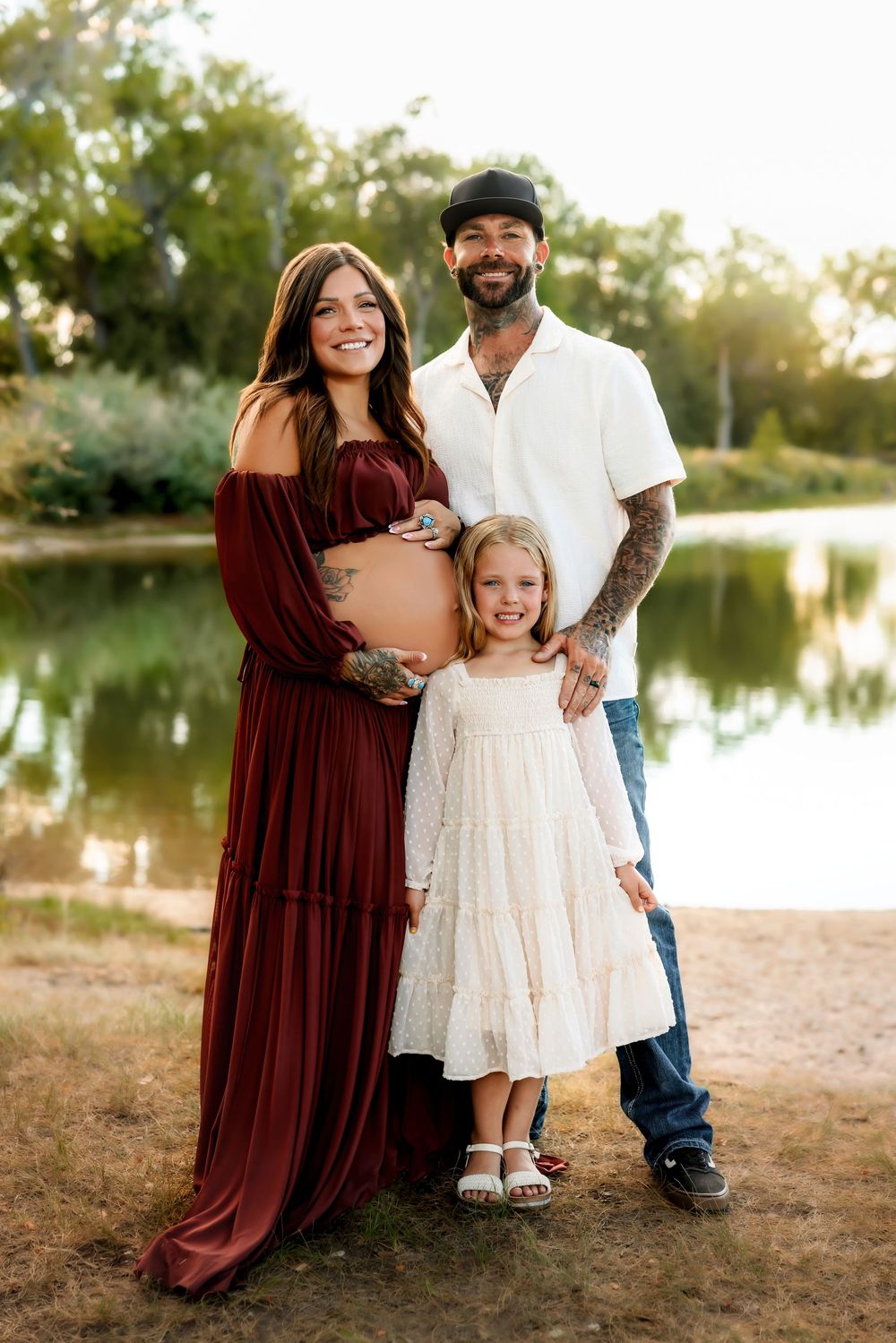A family in coordinating burgundy and white outfits poses together on a beach at sunset near a serene lake.