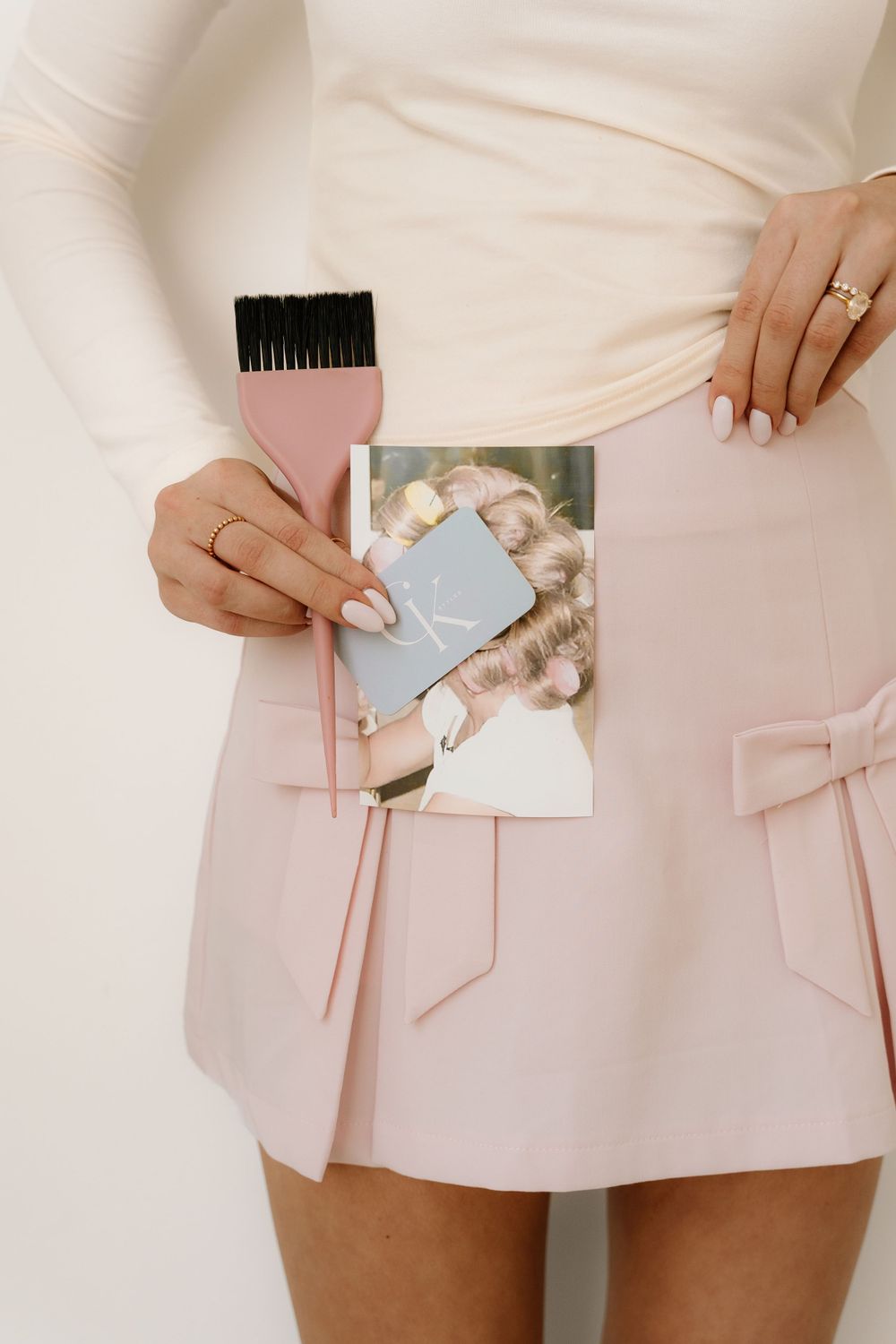 Hands holding a photograph and pink makeup brush against a white top and pink skirt background.