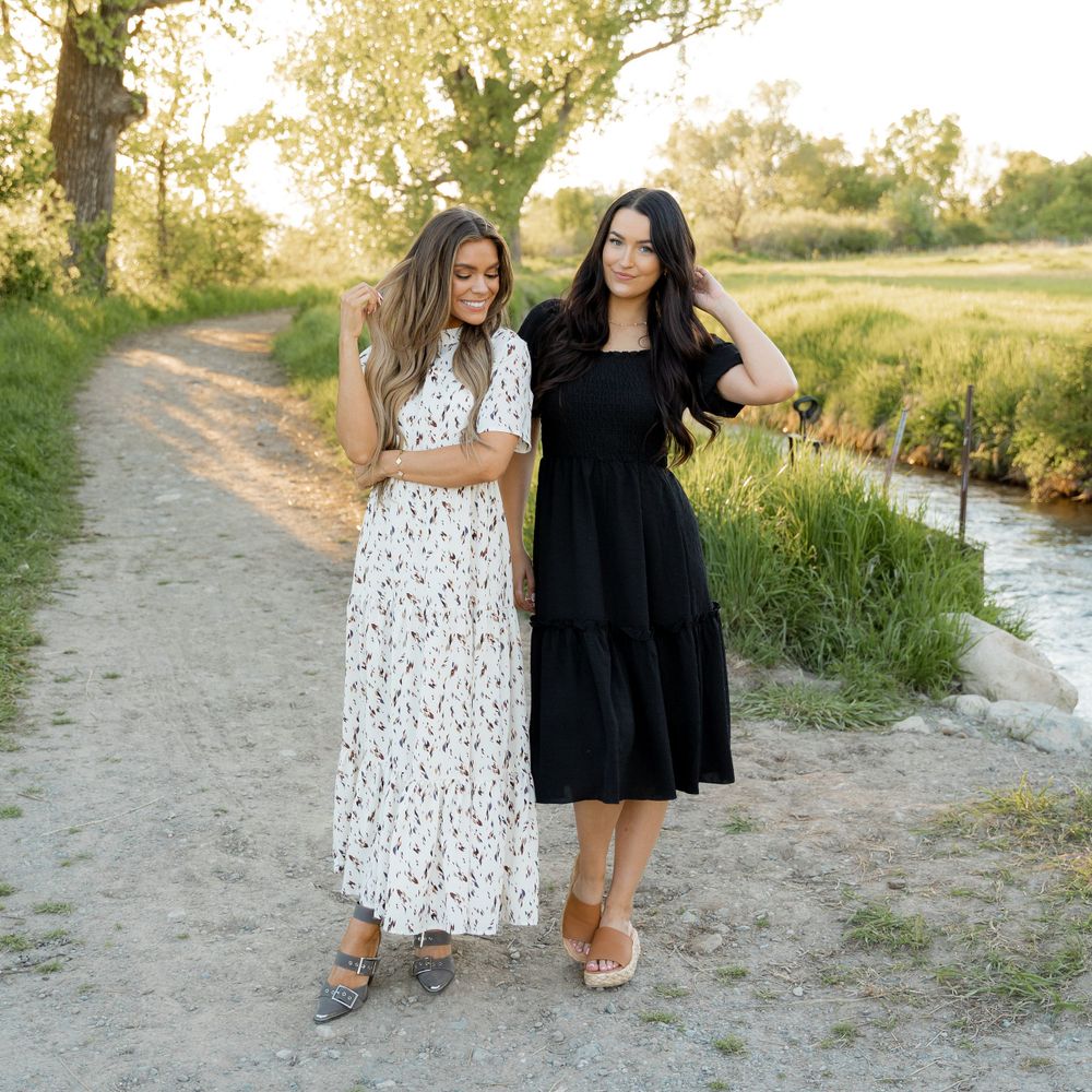 Two women in contrasting black and white dresses walking along rural stream path.