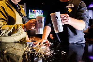 Hands pouring liquid into a cocktail shaker at a dimly lit bar while preparing drinks for service.