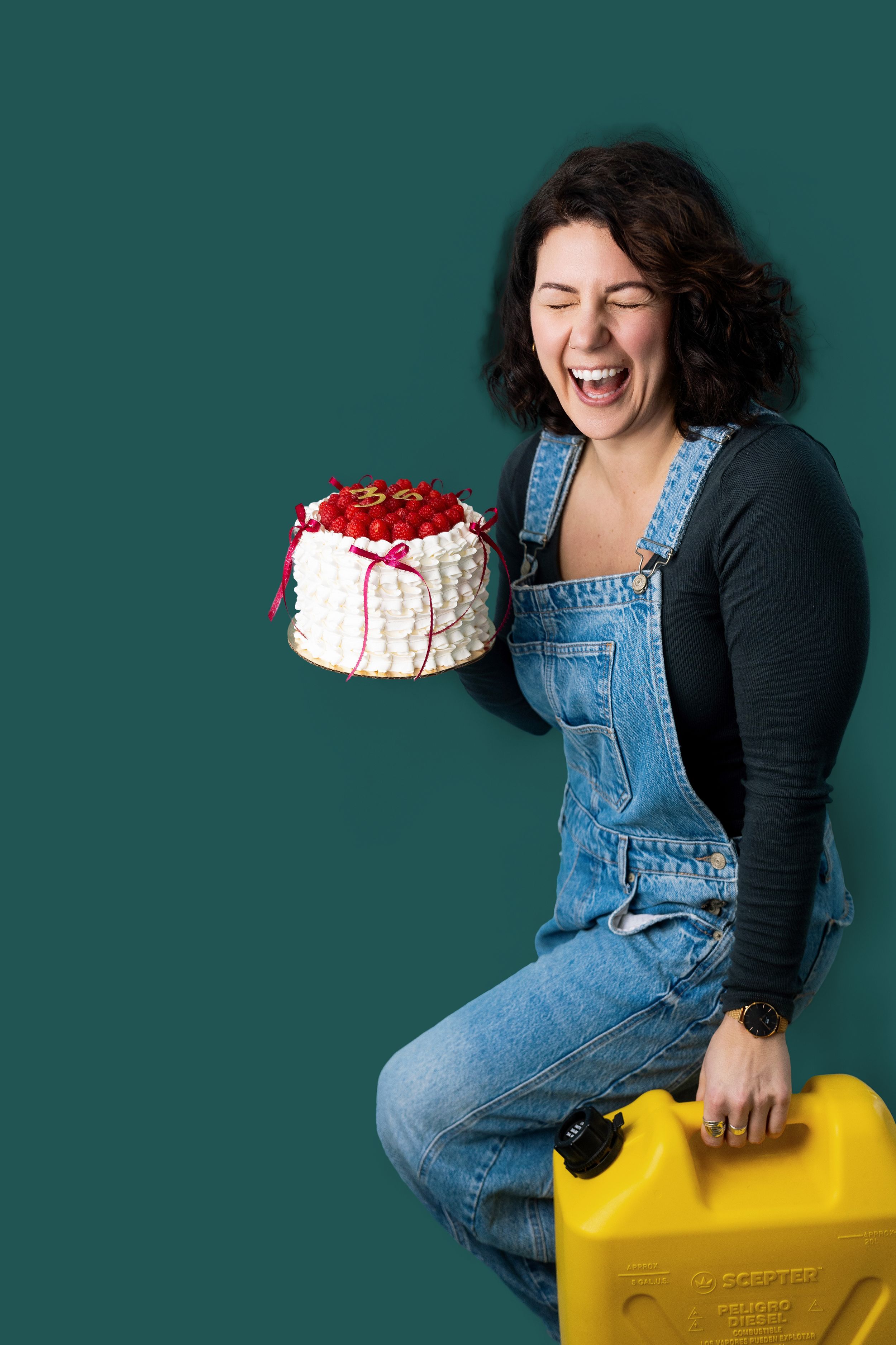 Person in denim overalls sits on yellow storage container while holding a red and white cake against teal background.