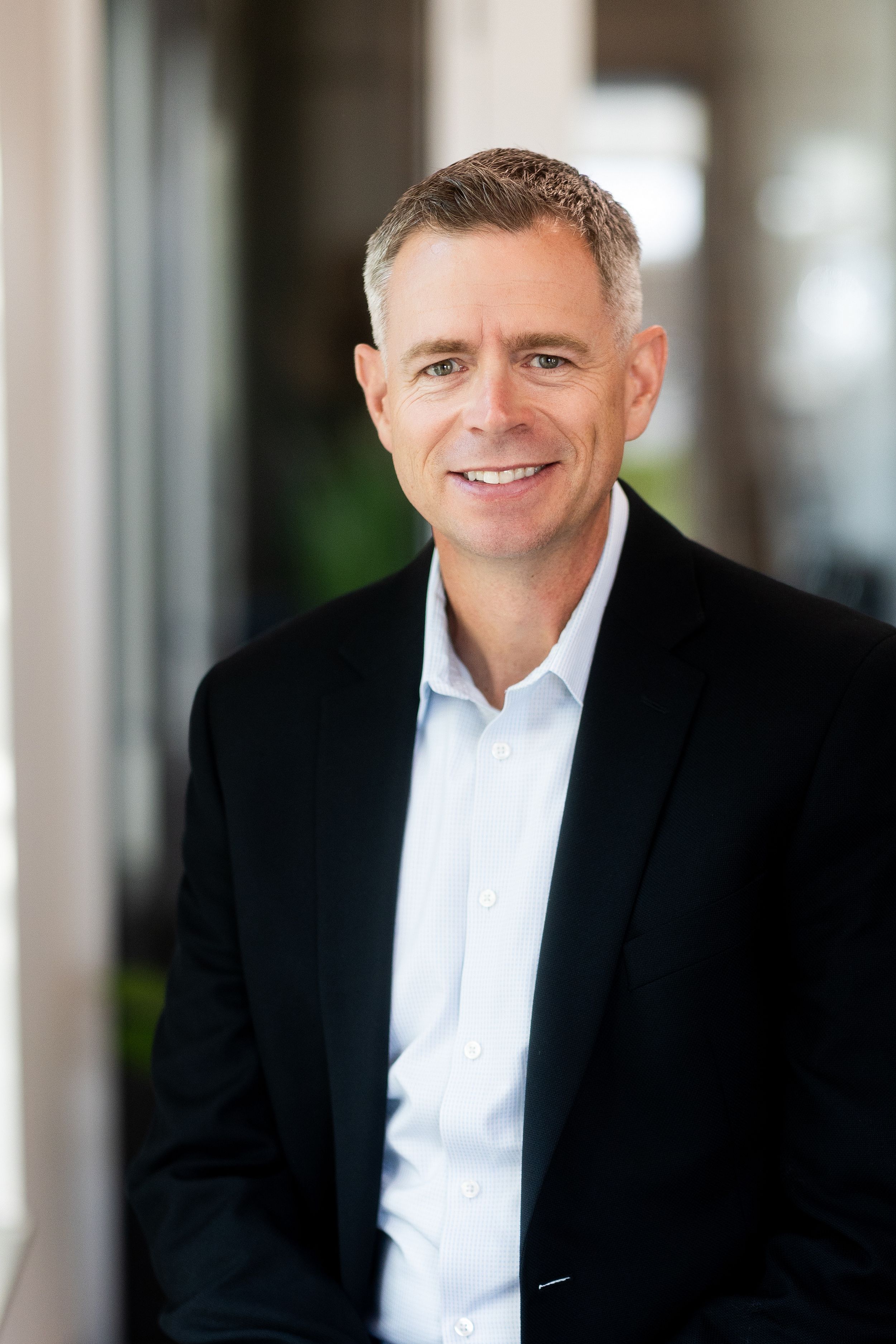 Professional headshot of someone in a black suit jacket and white dress shirt against a blurred office background.