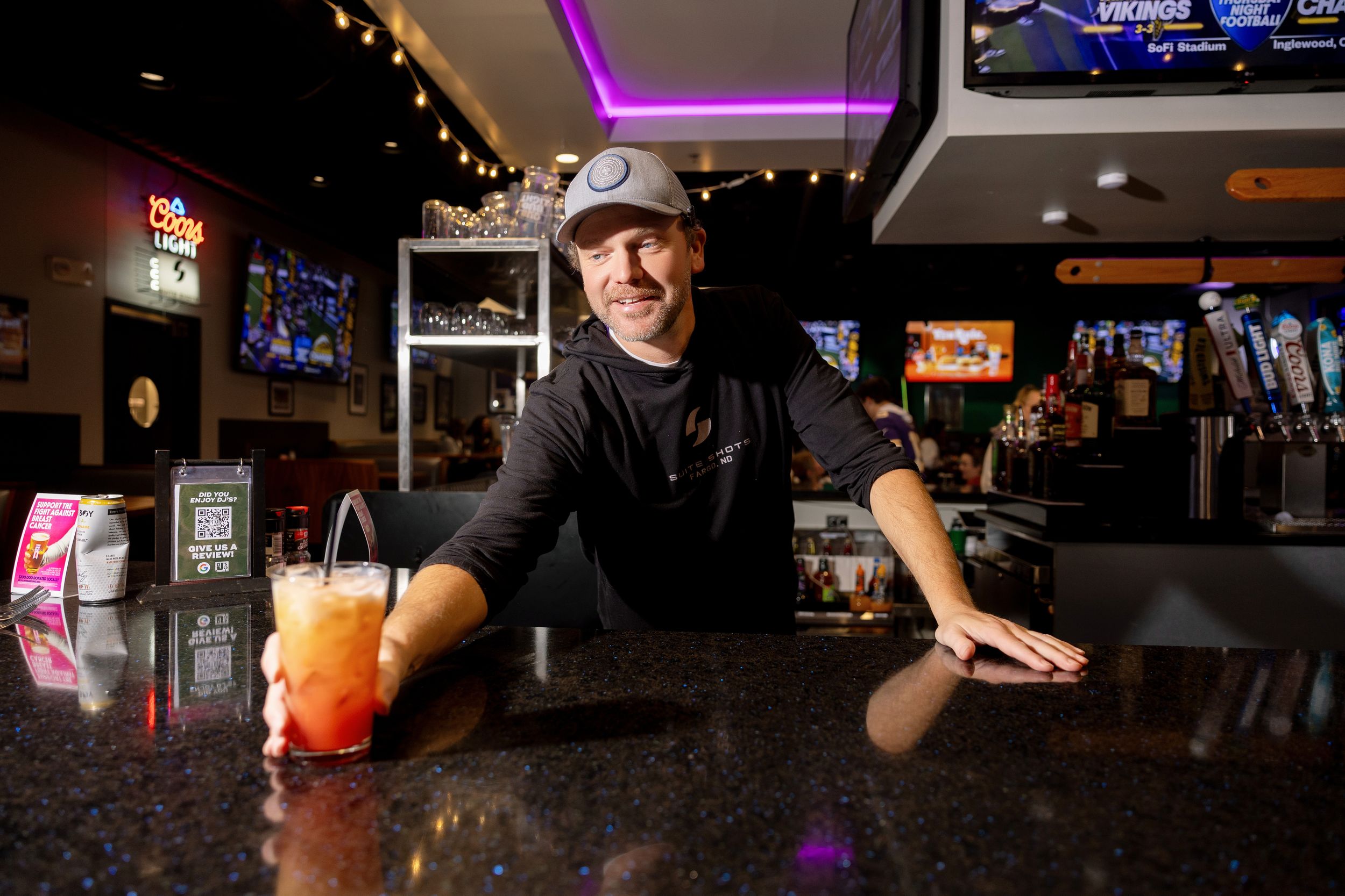 A person serves a bright orange cocktail at a dimly lit bar with neon lighting in the background.