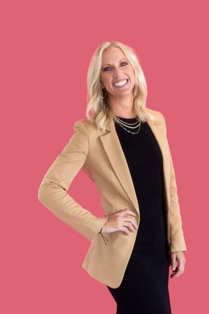 Professional studio portrait against pink background wearing black dress and beige blazer with bright joyful expression.