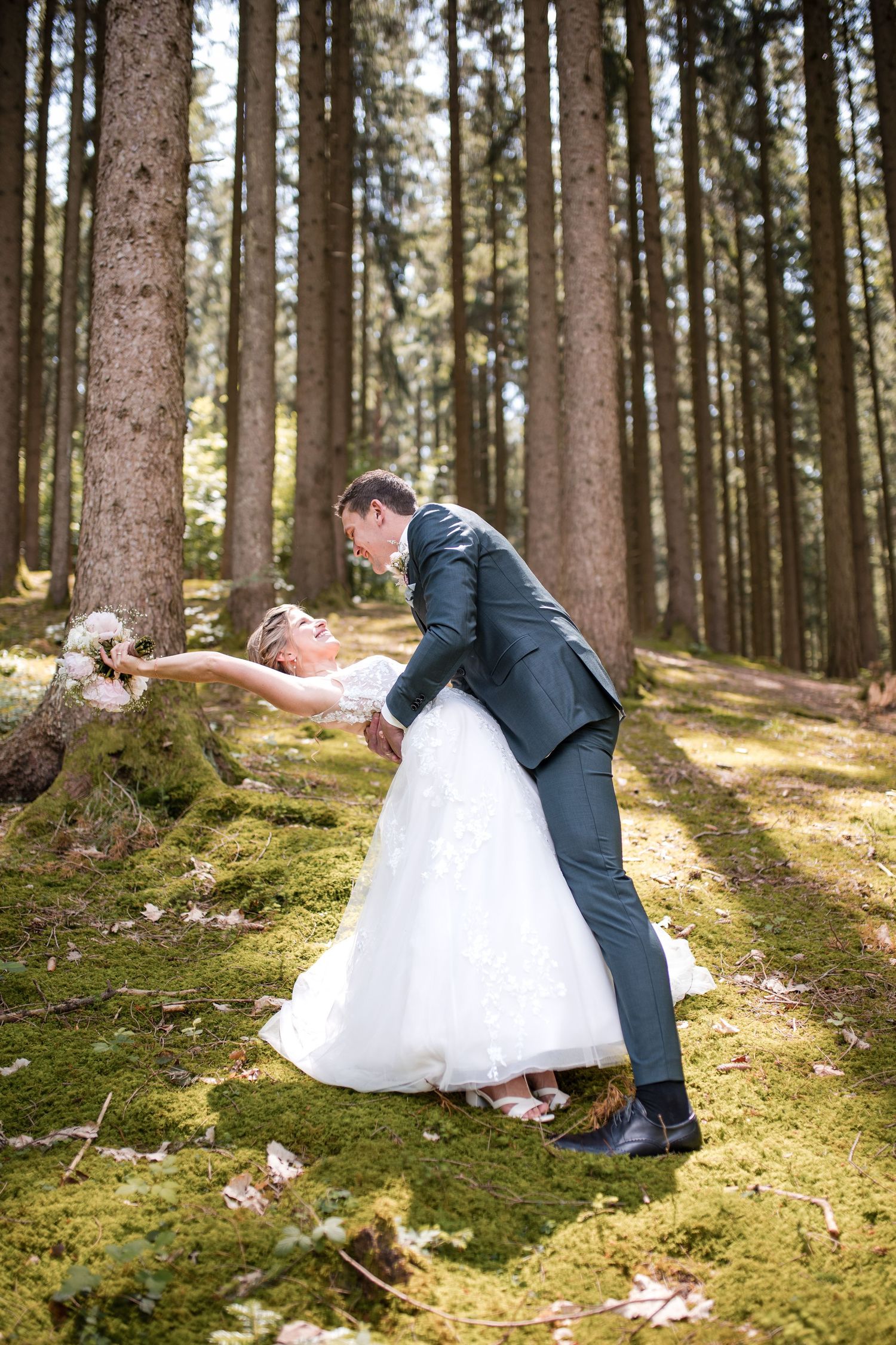 Romantic dip pose between newlyweds in a scenic pine forest with moss-covered ground.